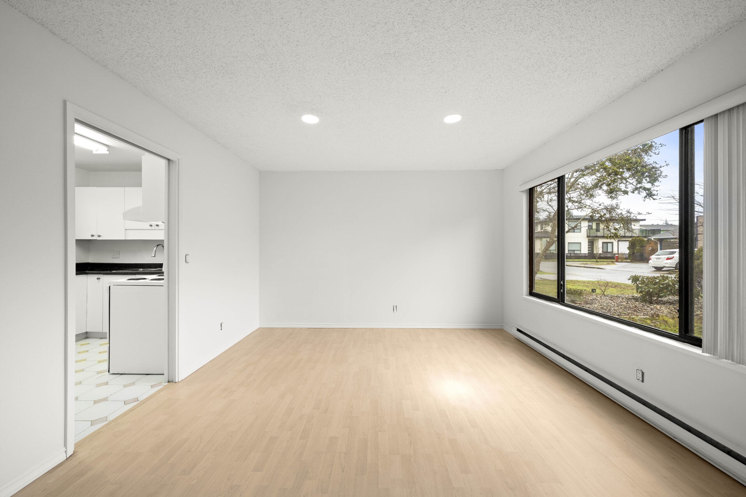 Unfurnished room with light wood flooring, large window, white walls, and view into a white kitchen. Natural light enters through the window facing a residential street.