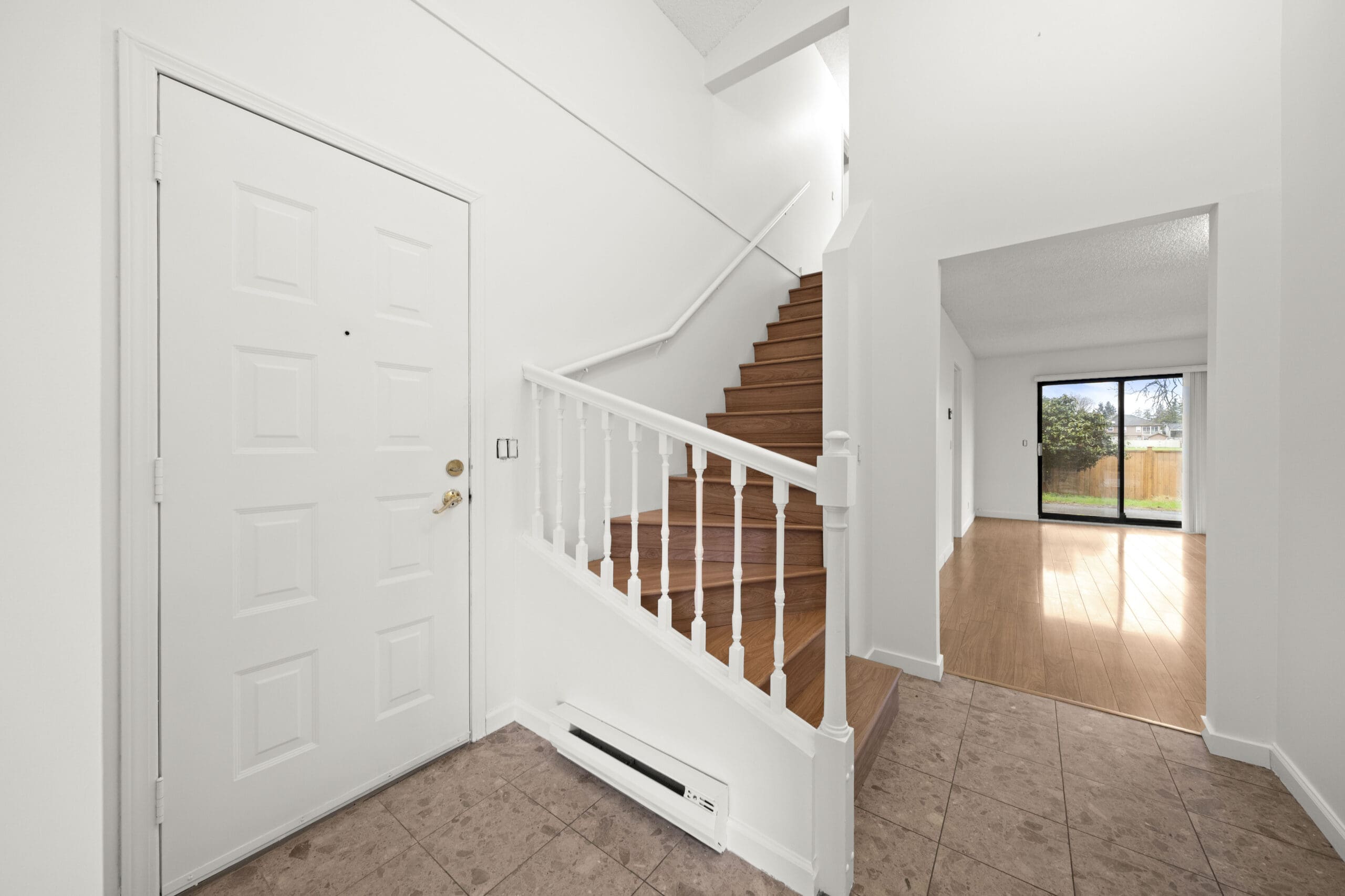 A residential entryway with a white front door, tiled floor, wooden staircase with white railing, and a view into a living area with wood flooring and sliding glass doors.