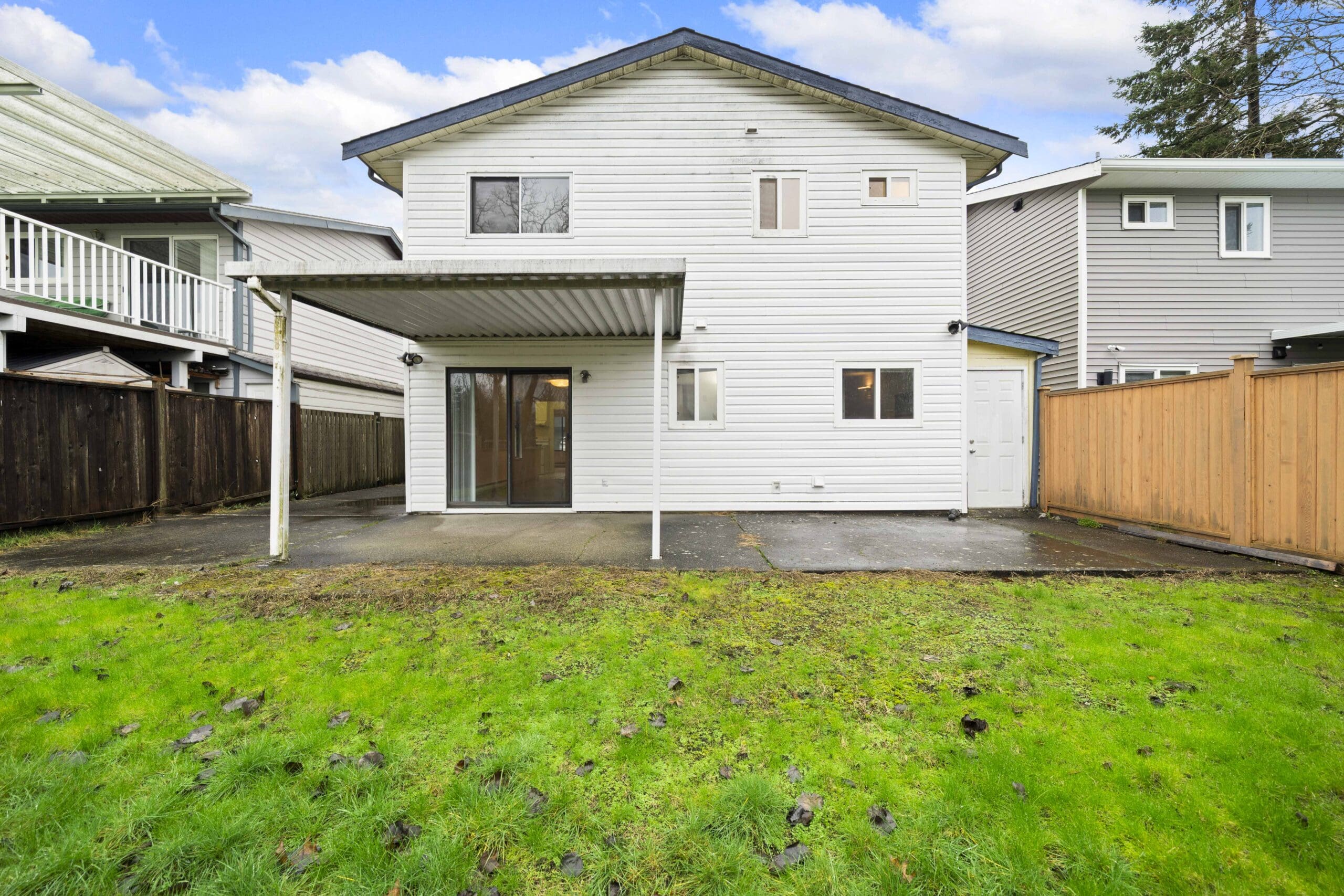 A two-story white house with a covered patio, sliding glass doors, and a fenced backyard with green grass and a paved area.