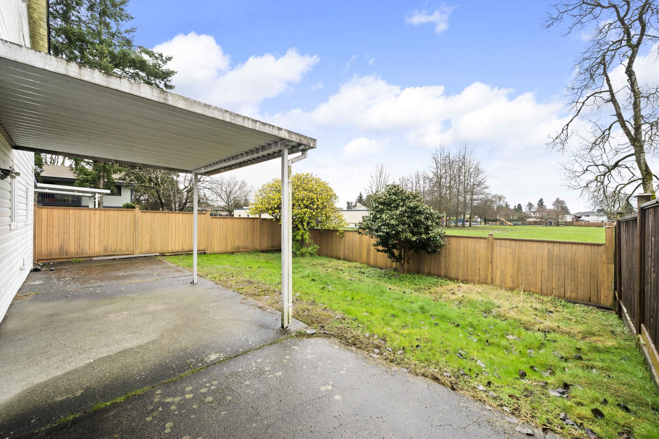 A backyard with a covered concrete patio, grassy area, wooden fence, and trees, under a partly cloudy sky.