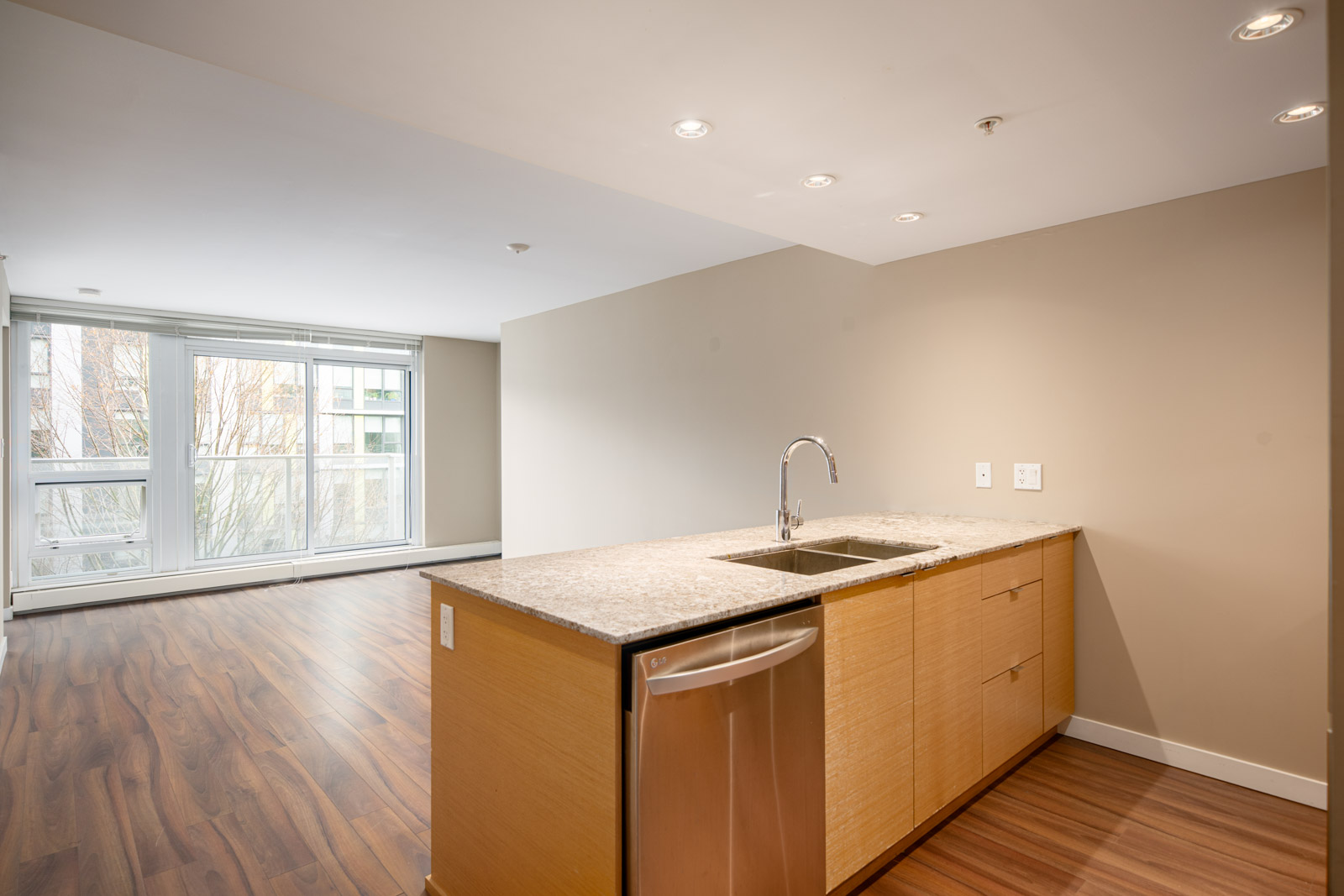 Modern open-plan apartment interior with a kitchen island, stainless steel sink, wood flooring, and large windows providing natural light.