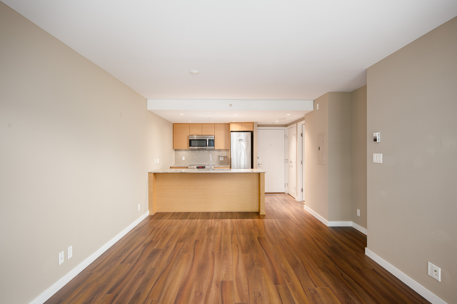 Unfurnished apartment interior with wood floors, beige walls, and an open kitchen featuring light wood cabinets and stainless steel appliances.