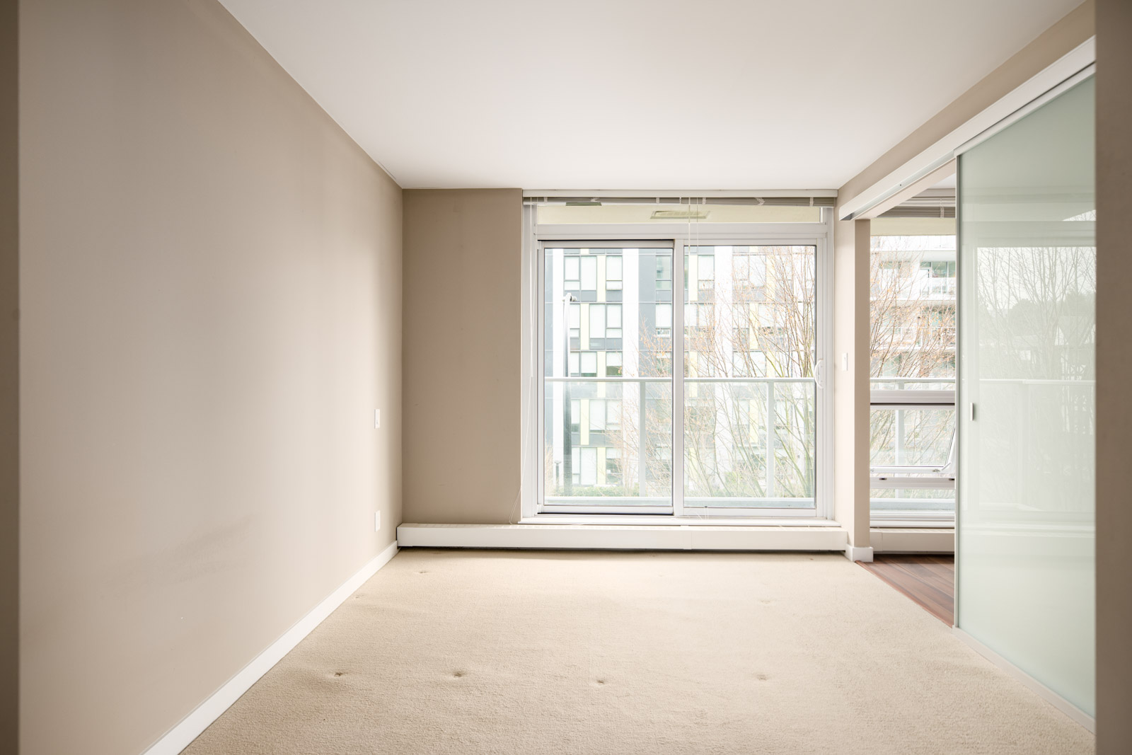 Empty room with beige walls and carpet, large window and glass sliding door, overlooking a modern building and trees outside.