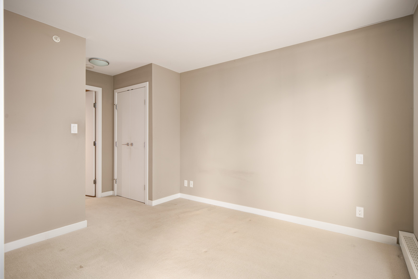 Empty room with beige carpet and walls, a white closet door, and a doorway leading to a hallway. The room is well-lit with natural light.