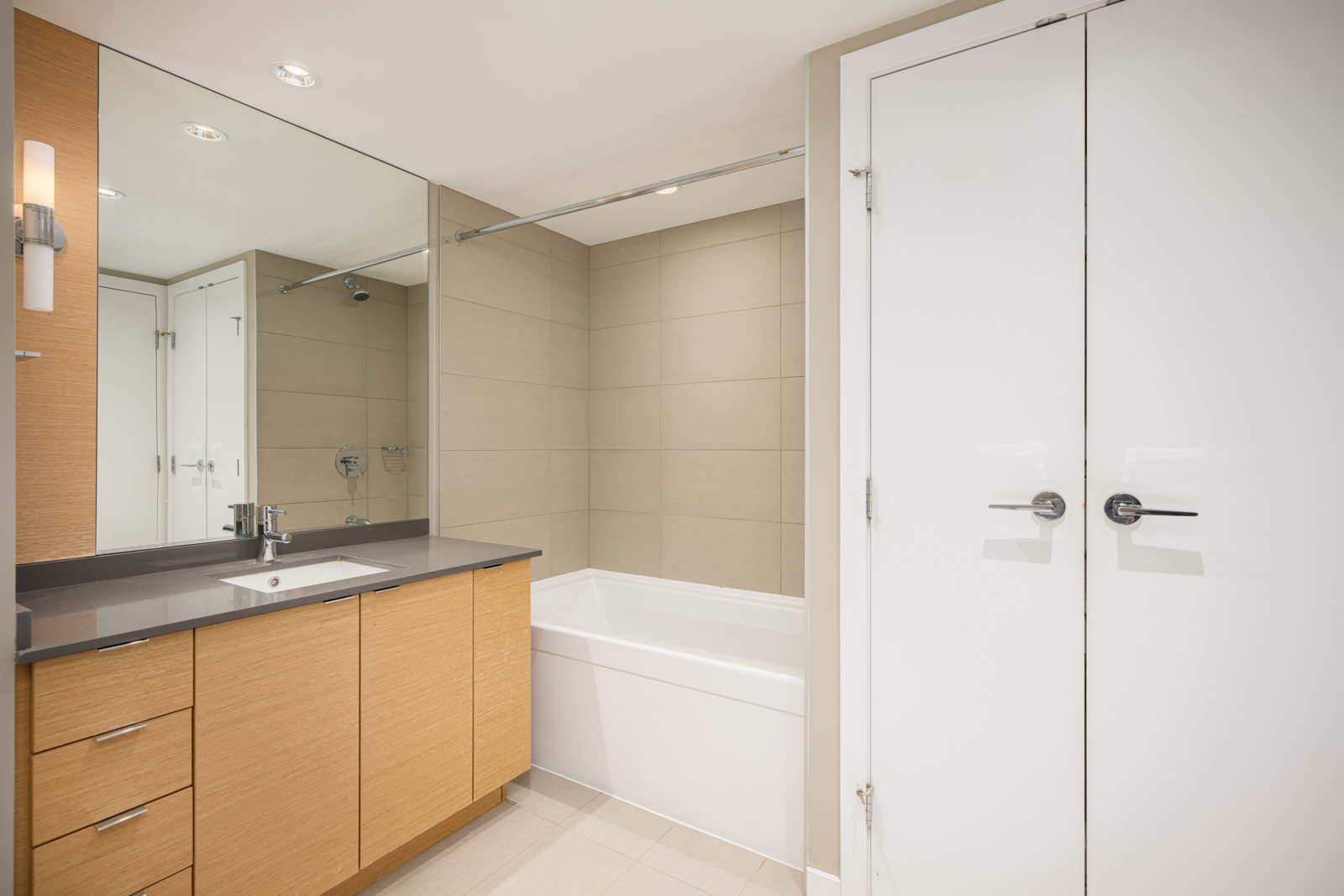 Modern bathroom with a wooden vanity, black countertop, large mirror, bathtub with beige tile surround, and white double-door closet.