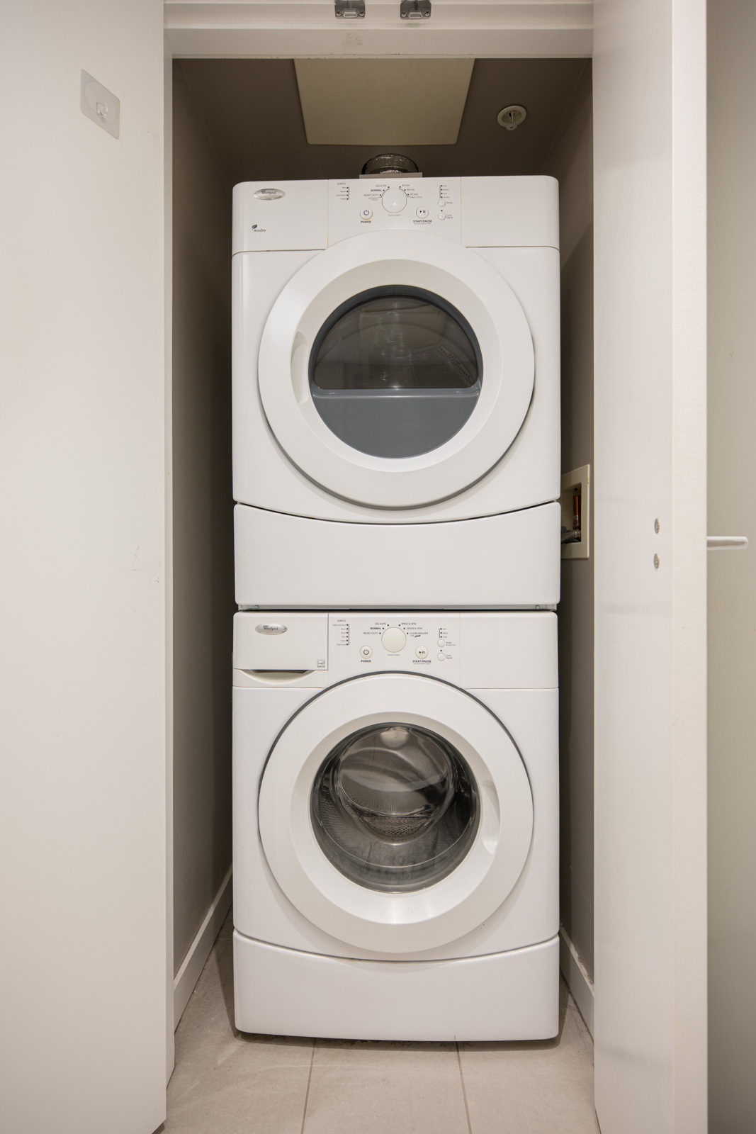 A stacked washer and dryer unit is placed in a small laundry closet with white walls and a tiled floor.