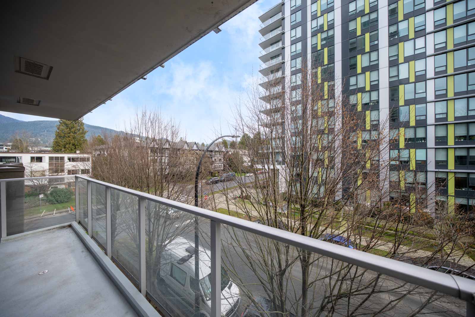 View from a balcony overlooking a street, trees, and a tall modern apartment building with green accents; mountains are visible in the background.