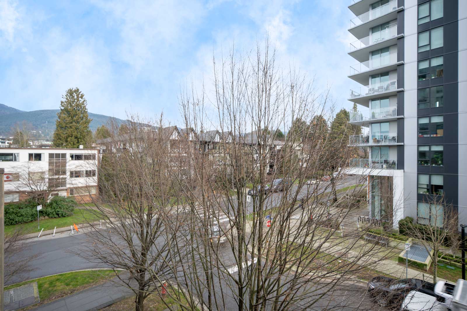 View of a street corner with leafless trees, a modern apartment building on the right, and other buildings in the background on a clear day.