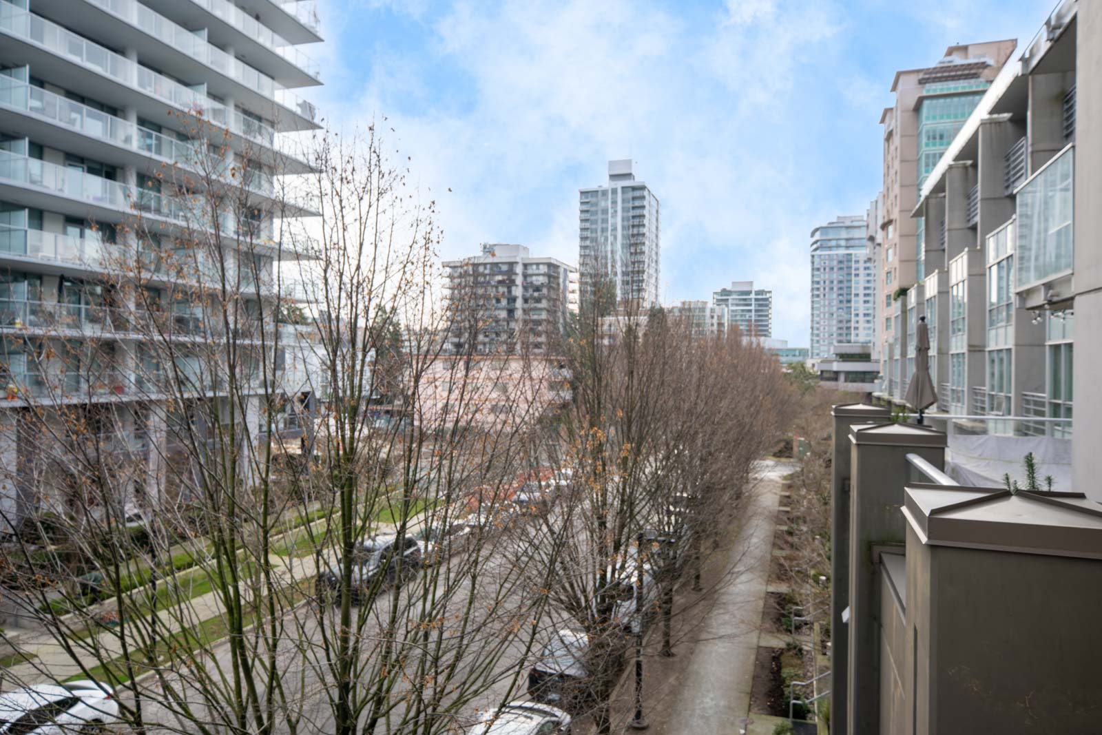 A city street lined with leafless trees, flanked by modern mid-rise apartment buildings under a partly cloudy sky.