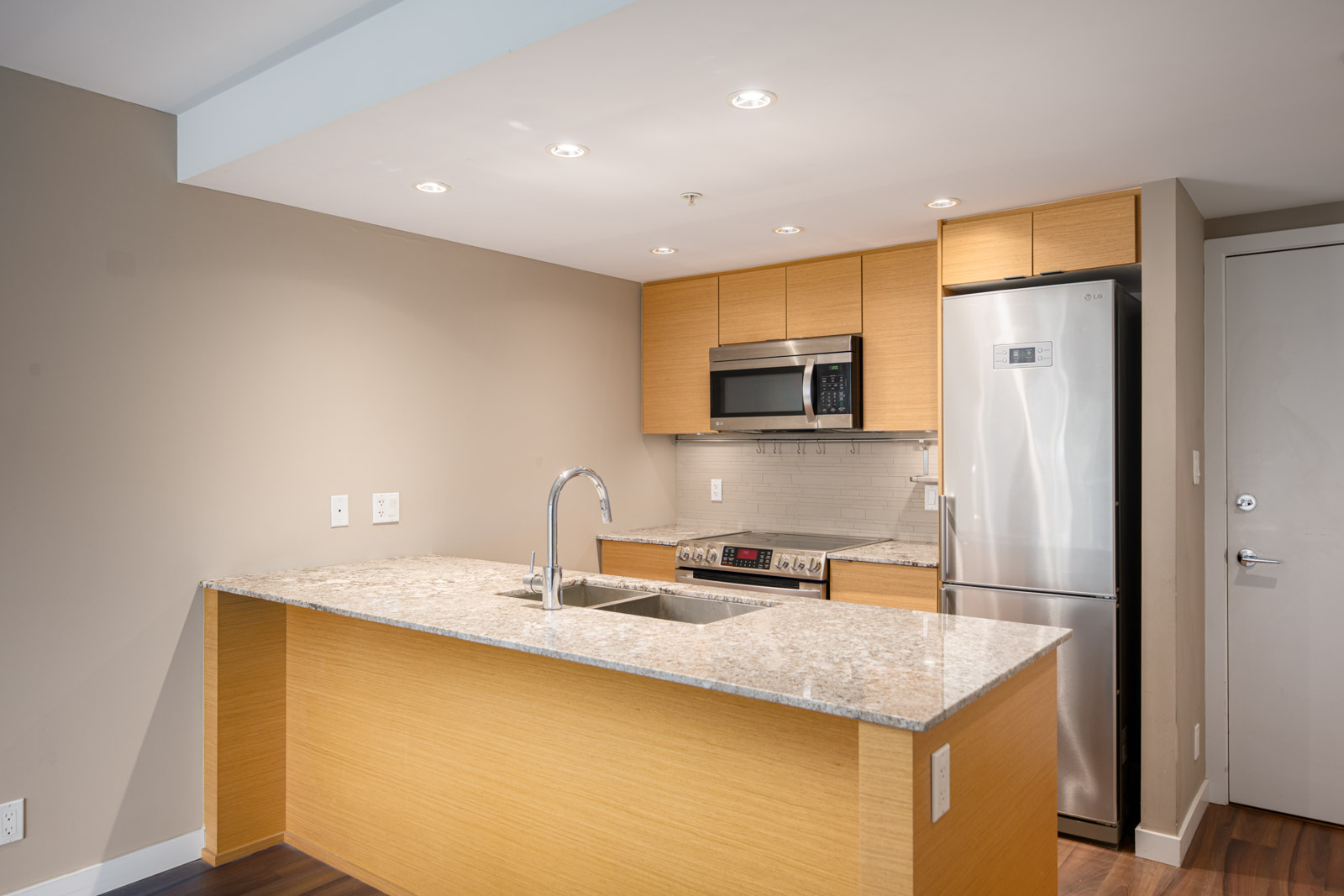 Modern kitchen with light wood cabinets, granite countertop island with sink, stainless steel appliances, and recessed lighting.