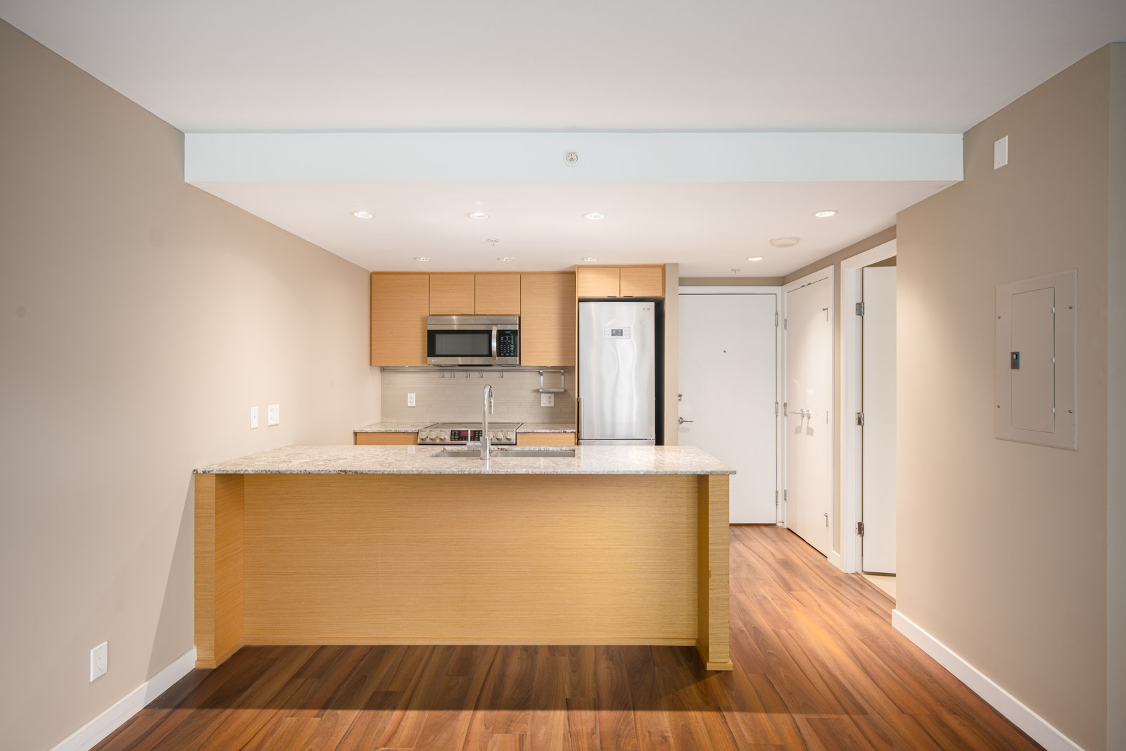 Modern kitchen with wood cabinets, granite countertop, stainless steel appliances, and hardwood flooring in a neutral-toned open layout.