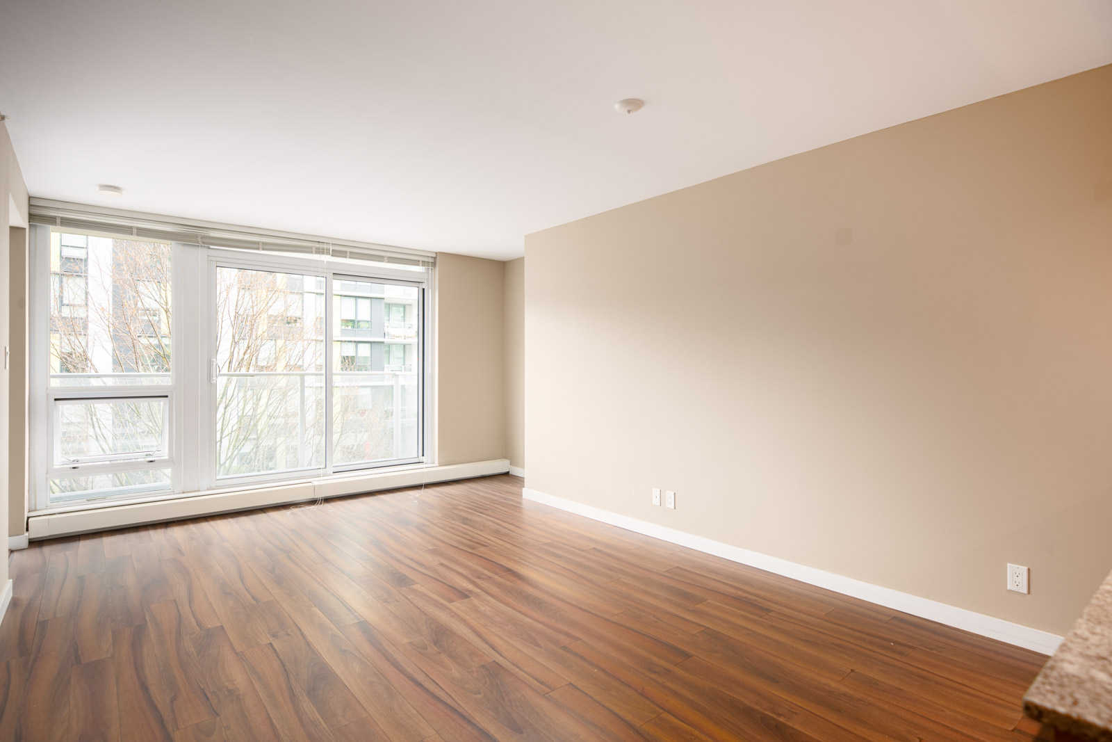 Unfurnished room with large window, beige walls, and wood flooring; daylight streams in from outside.