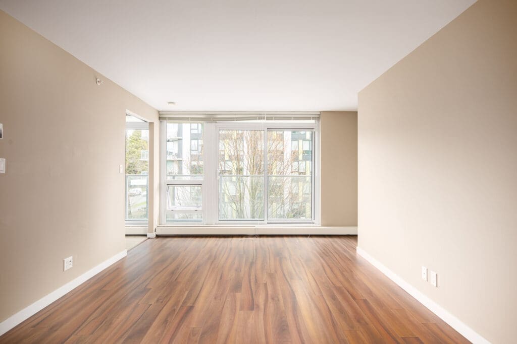 Empty room with beige walls, large windows, and wood flooring. Natural light enters from the windows overlooking trees and buildings outside.