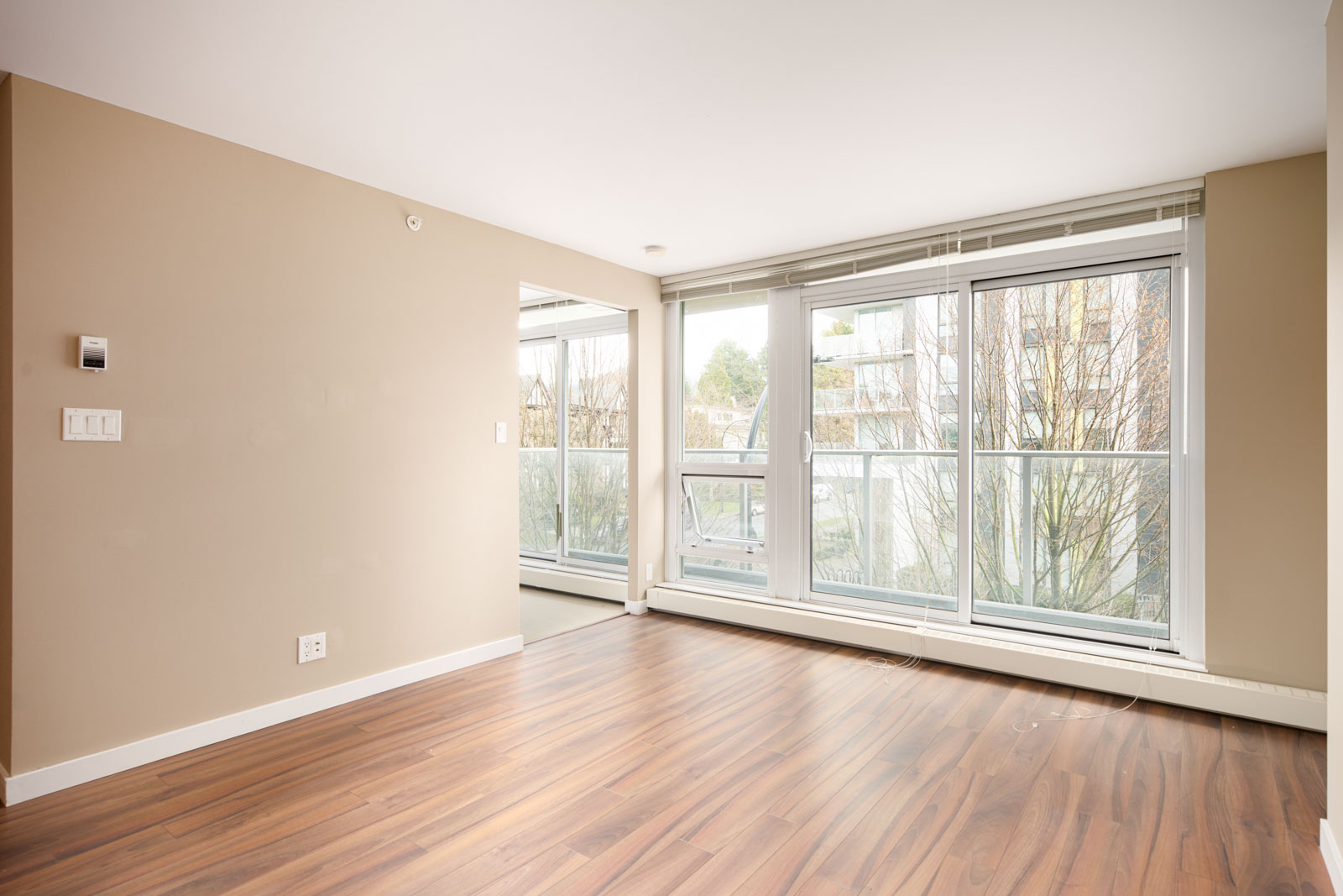 Unfurnished room with beige walls, wood flooring, and large floor-to-ceiling windows overlooking trees and buildings.