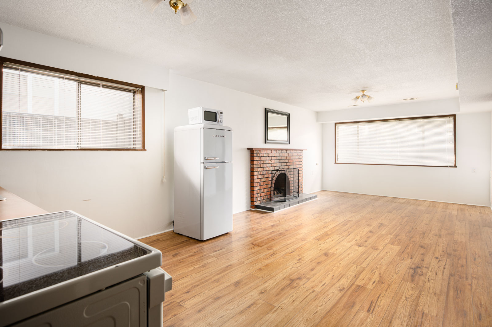 A bright, unfurnished room with wood flooring, large windows, a brick fireplace, a white fridge, a microwave, and a stove in the corner.