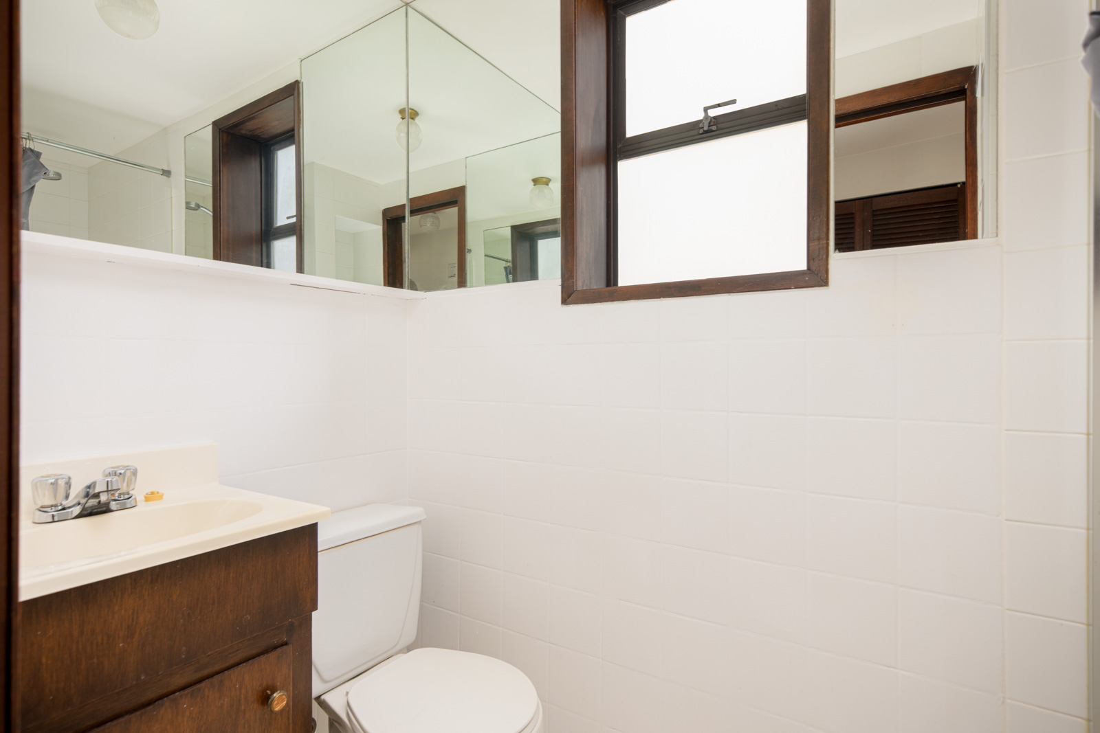 Small bathroom with white tiled walls, a toilet, a dark wood vanity with sink, a window, and large mirrors reflecting the space.