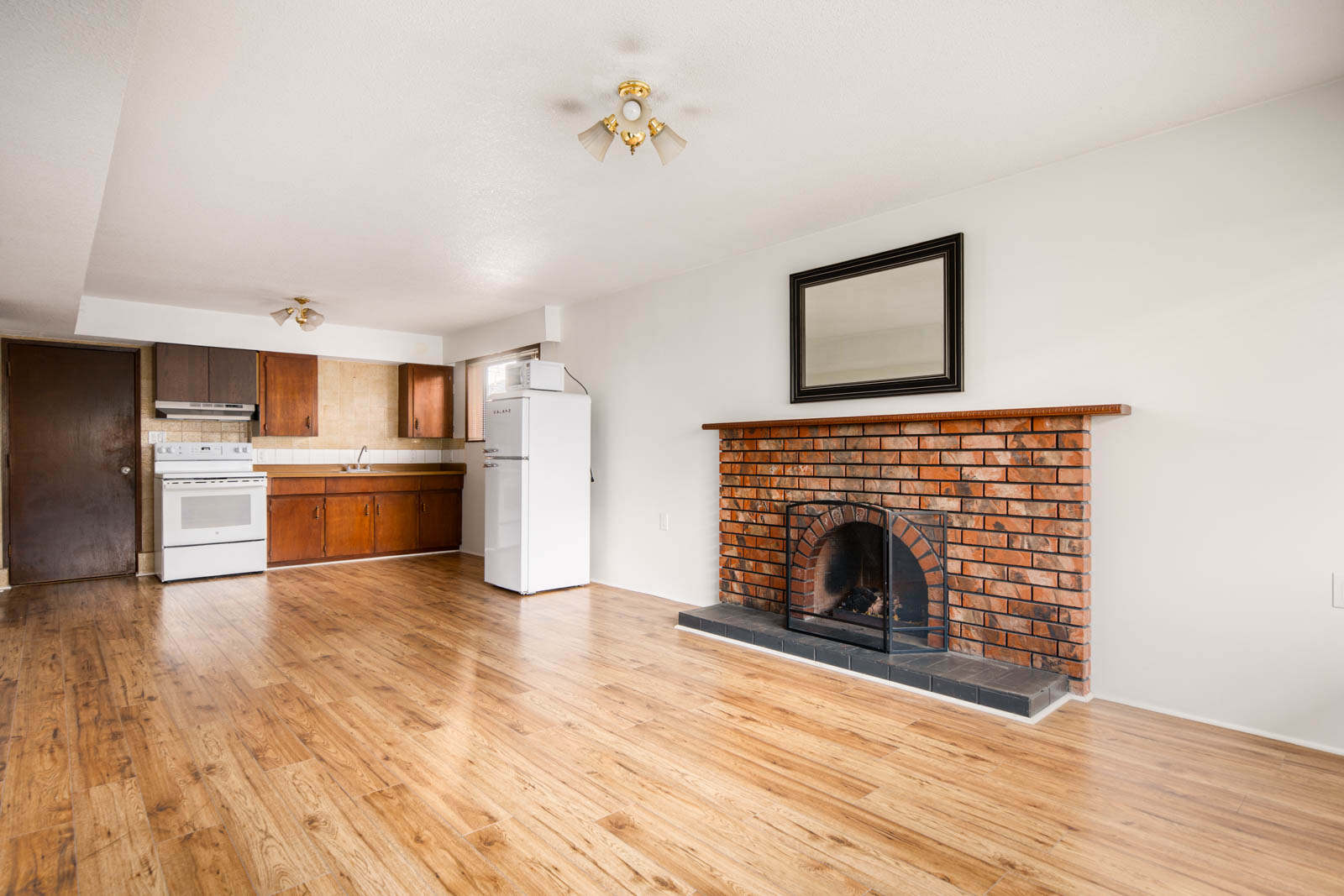 Unfurnished living area with wood flooring, brick fireplace, and adjacent kitchen featuring white appliances and brown cabinets.
