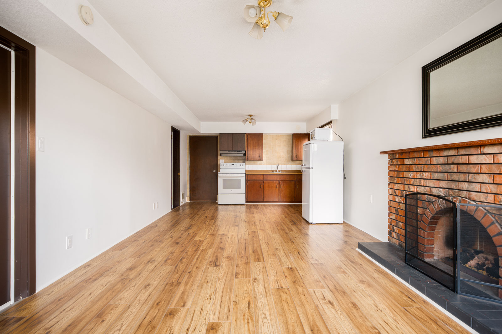 Unfurnished room with wood flooring, brick fireplace, and open kitchen area with white appliances and brown cabinets.