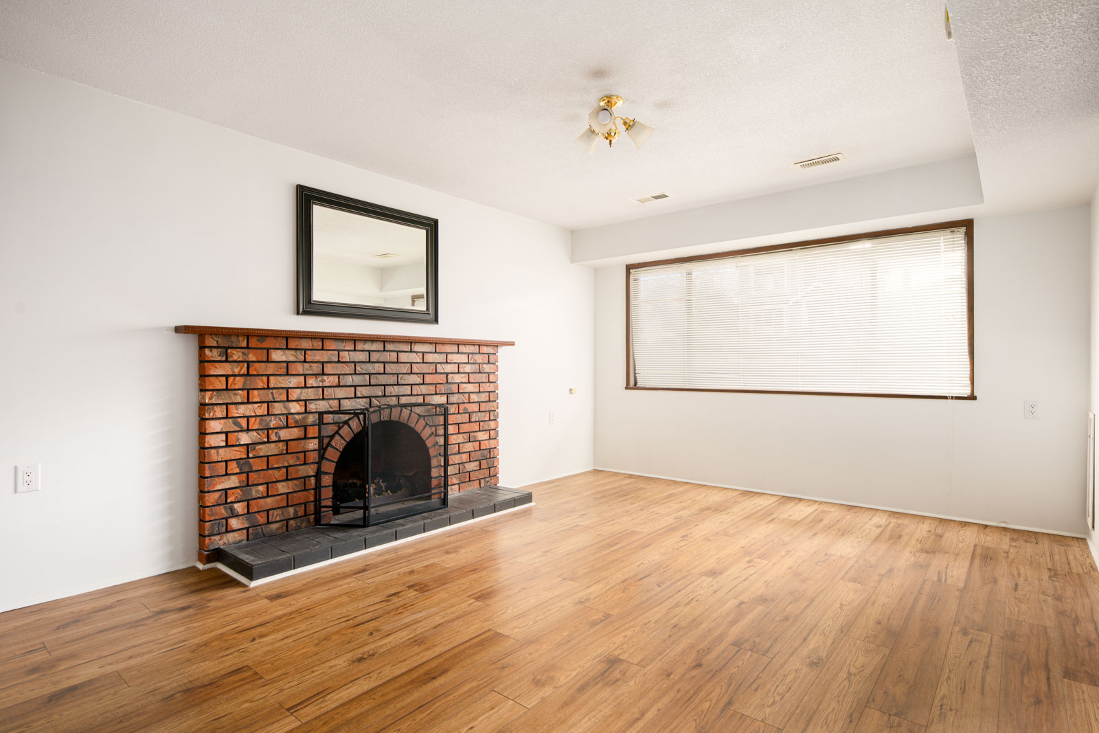 Unfurnished living room with wood flooring, a brick fireplace, a wall mirror above the mantel, and a large window with blinds.