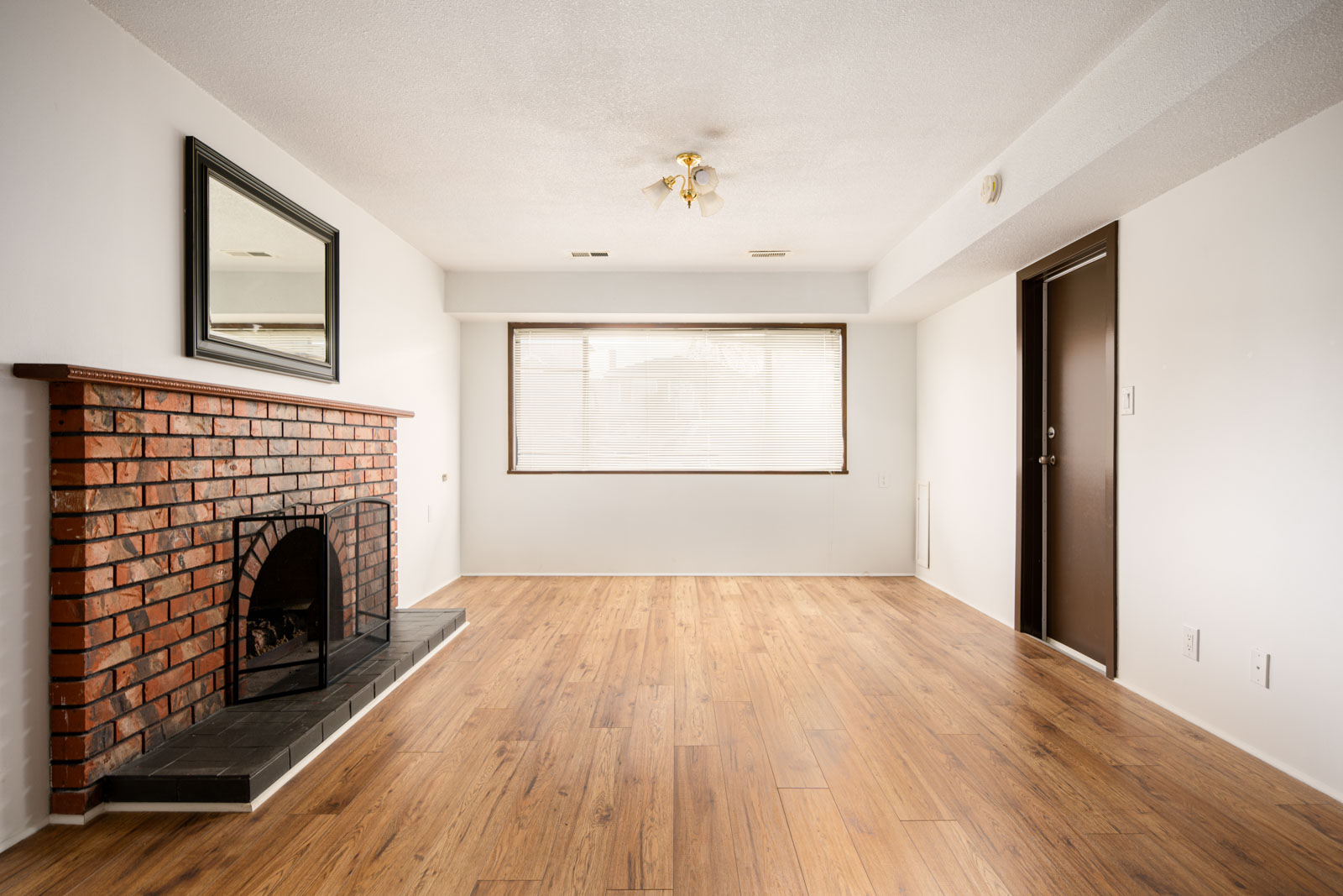 Empty living room with wood flooring, a red brick fireplace, a large window with blinds, white walls, and a brown door.
