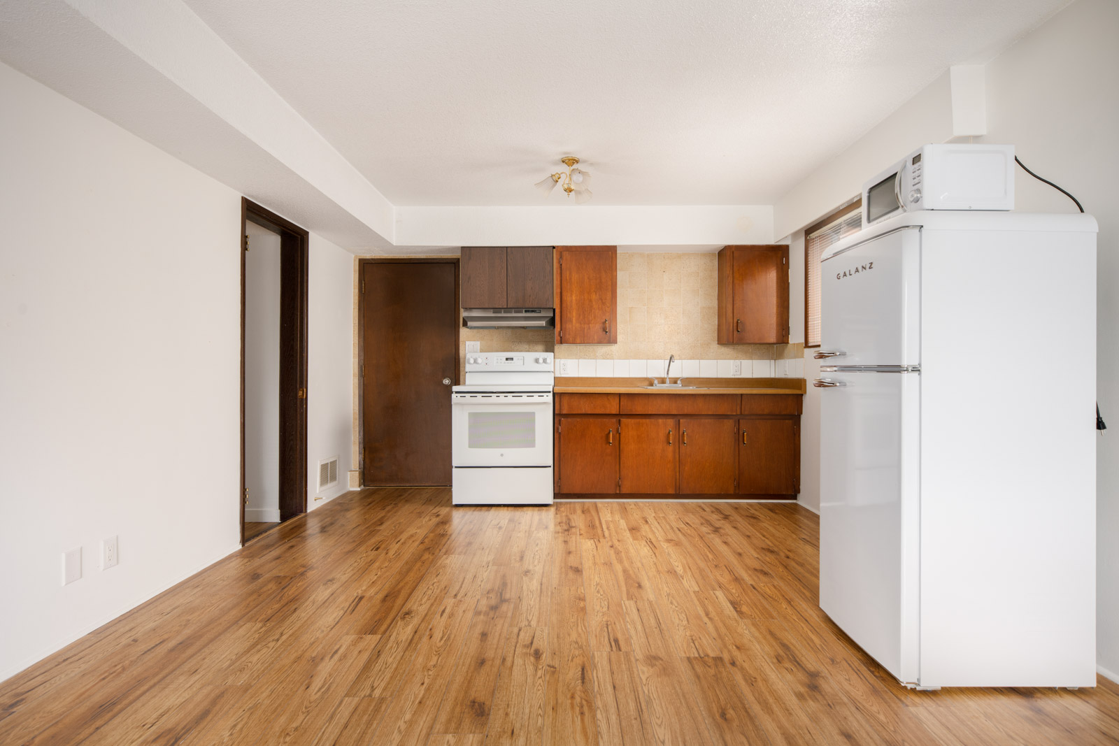 Unfurnished kitchen with wood cabinets, white stove and fridge, microwave on top, and wood laminate flooring in an empty room with white walls.