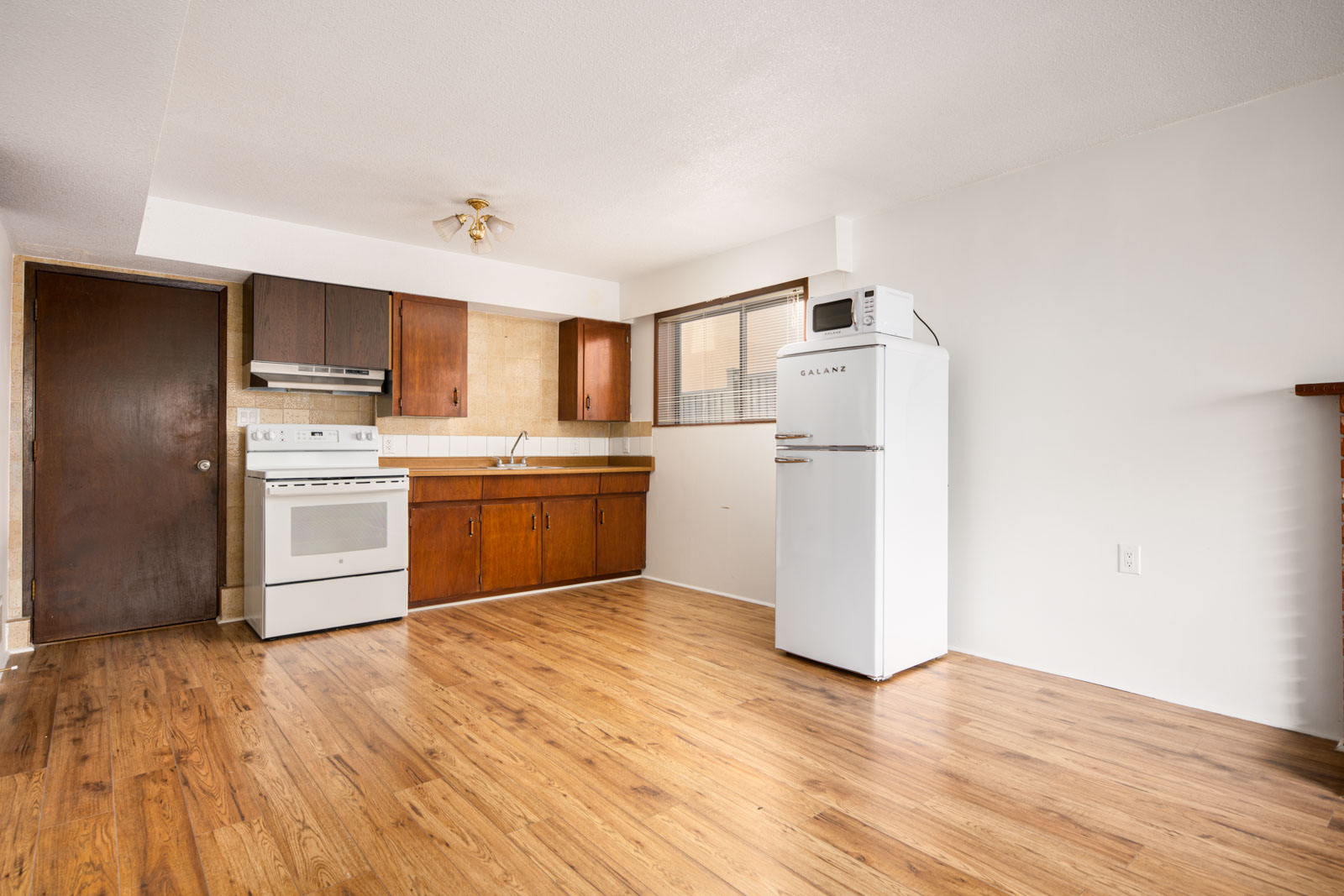 Small kitchen with wood cabinets, white stove, fridge, and microwave. The room has wood flooring, white walls, and a large window near the refrigerator.