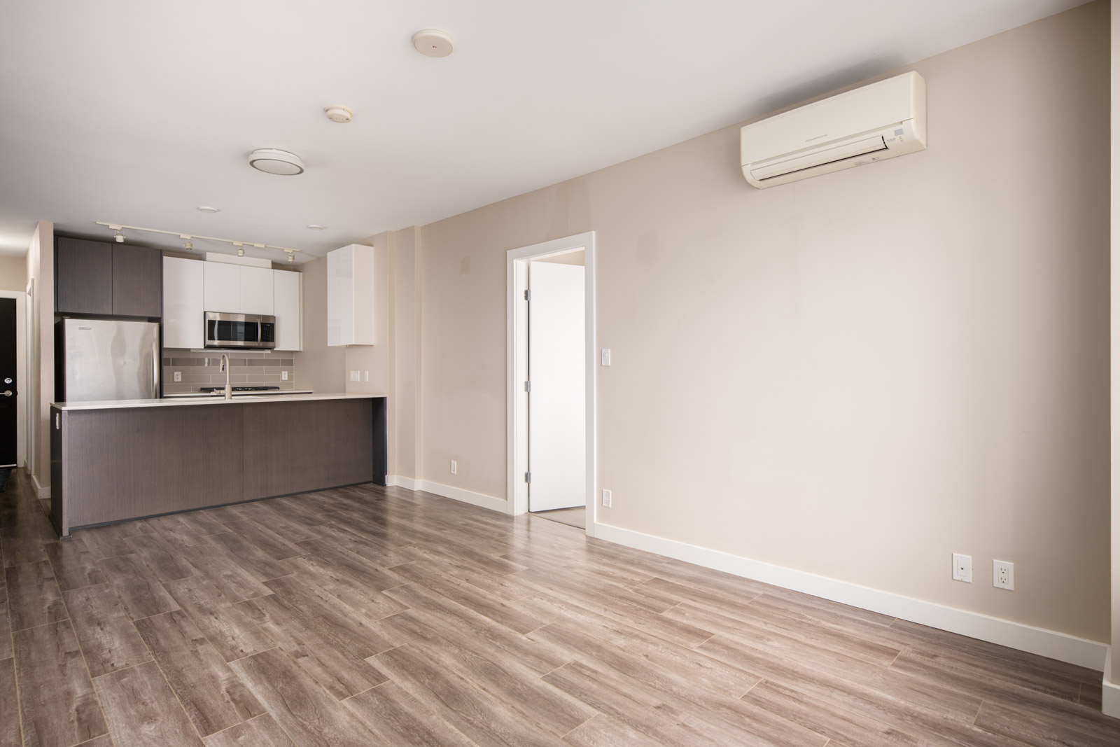 Modern empty apartment interior with wood flooring, beige walls, an open kitchen with stainless steel appliances, and a wall-mounted air conditioning unit.