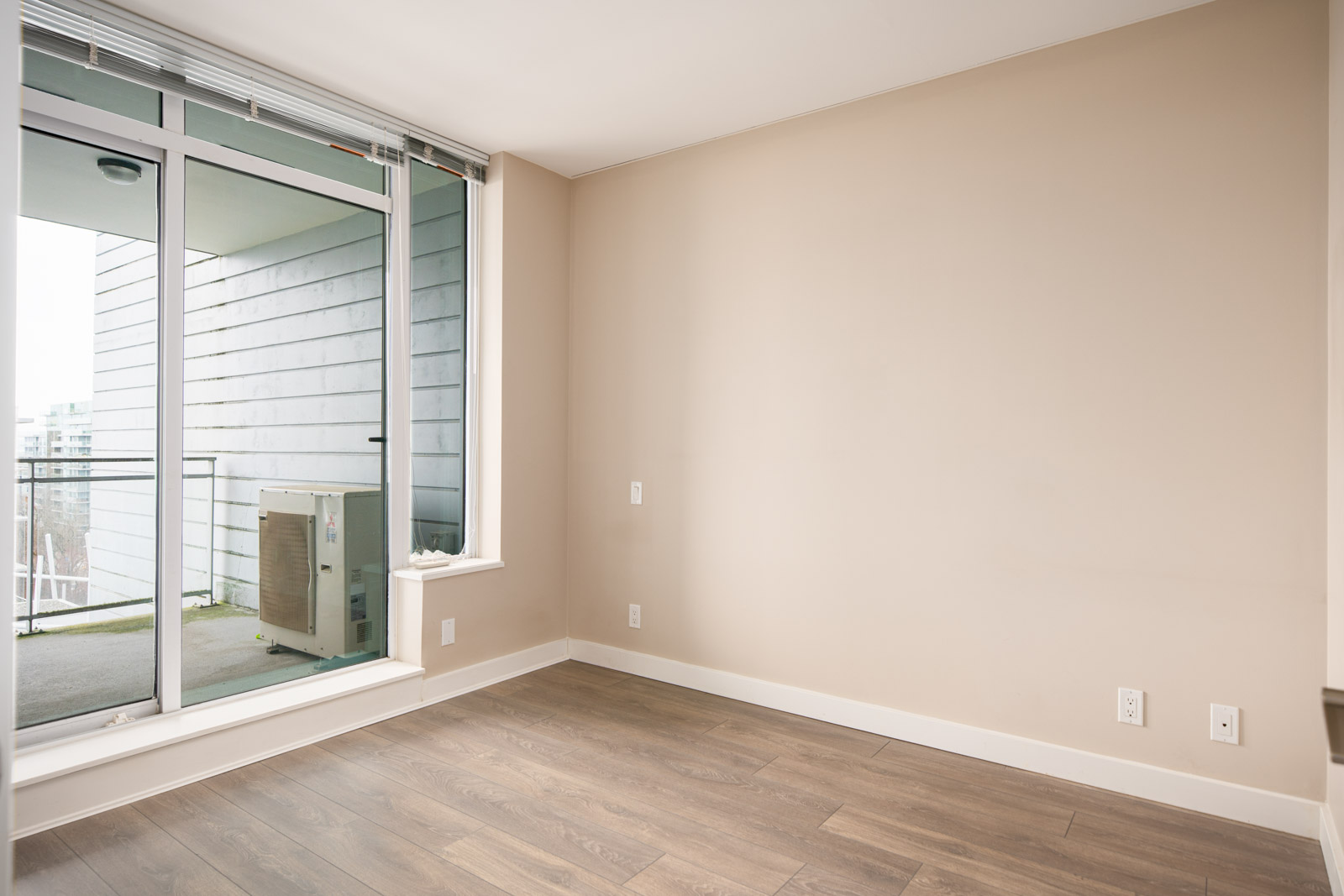 Empty room with beige walls, wood flooring, an electrical outlet, and a sliding glass door leading to an outdoor balcony with a view of neighboring buildings.