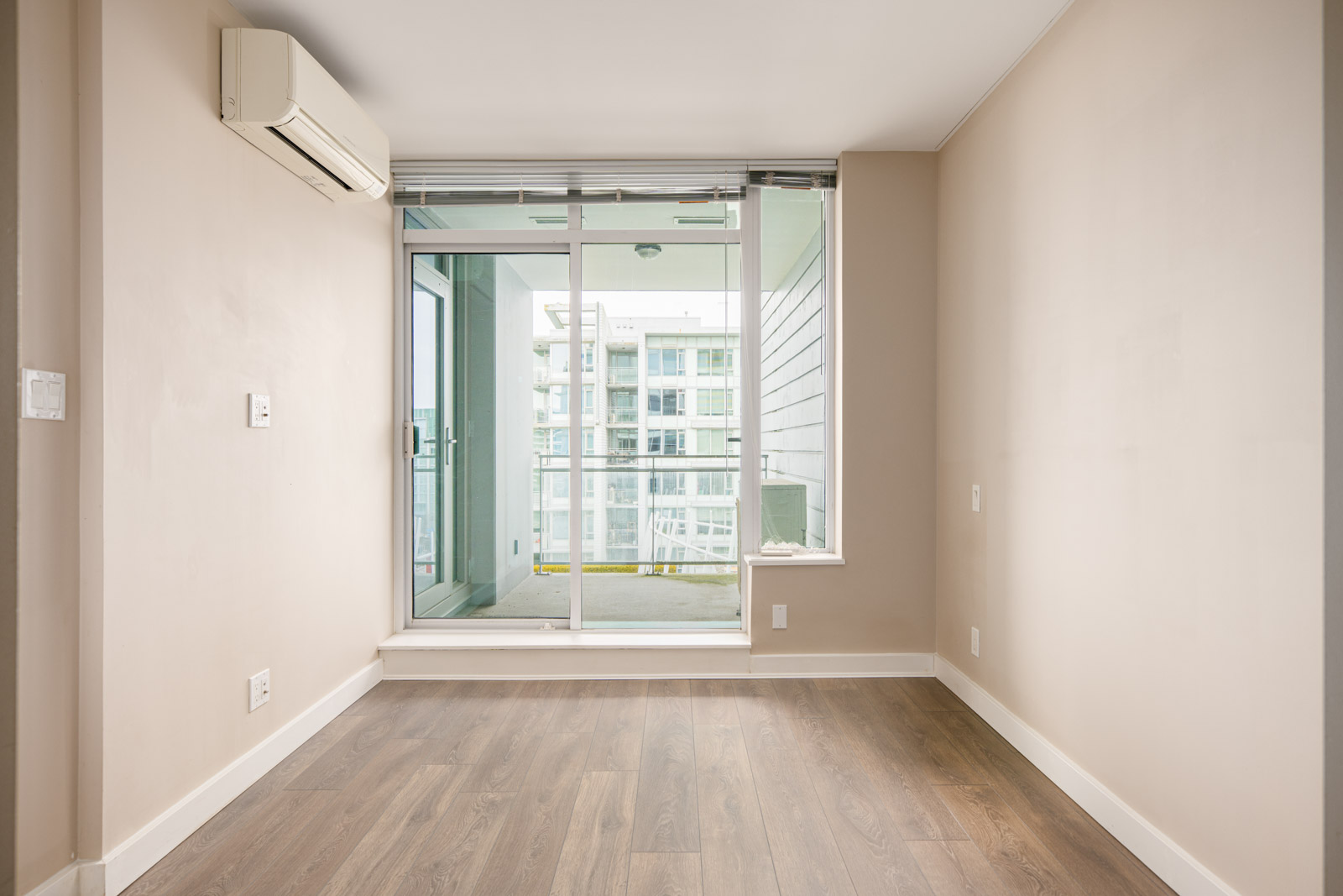 Empty room with beige walls, wood flooring, and a glass door leading to a balcony with a view of an apartment building. An air conditioning unit is mounted on the upper wall.