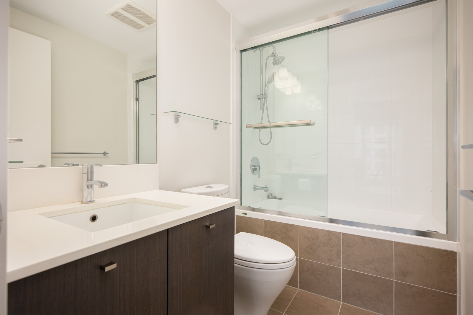Modern bathroom with a white sink, dark wood vanity, toilet, and bathtub with sliding glass doors and overhead shower. Light walls and brown floor tiles.