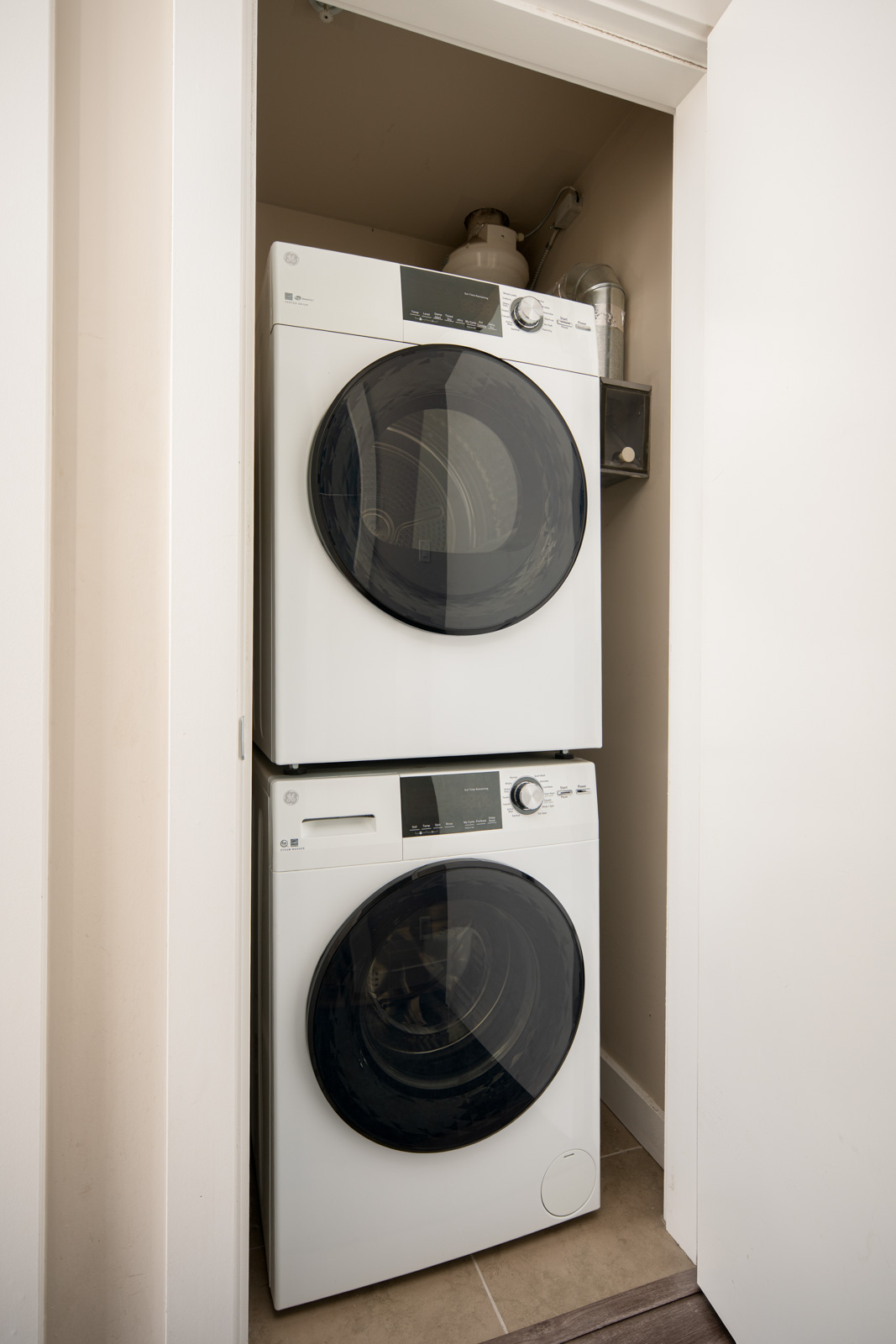 Stacked front-loading washer and dryer in a small laundry closet with beige walls and shelves holding cleaning supplies above the units.