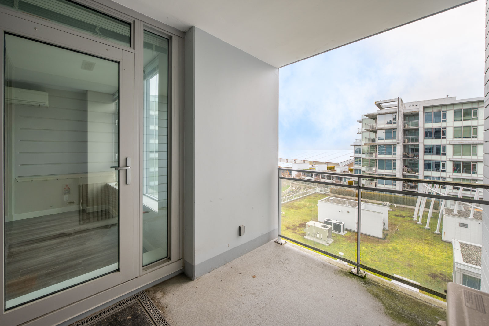 A concrete apartment balcony with glass railing overlooks a courtyard and neighboring modern apartment buildings under a cloudy sky.
