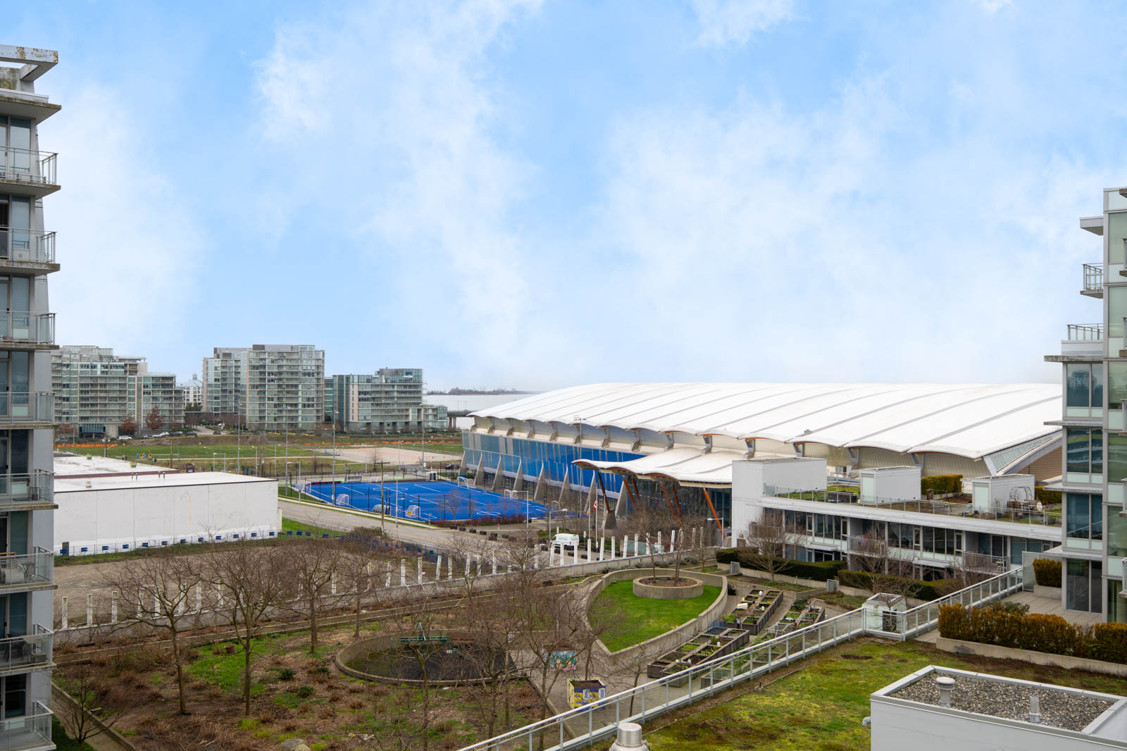 View of a large white-domed sports facility surrounded by apartment buildings, tennis courts, and landscaped green areas under a partly cloudy sky.
