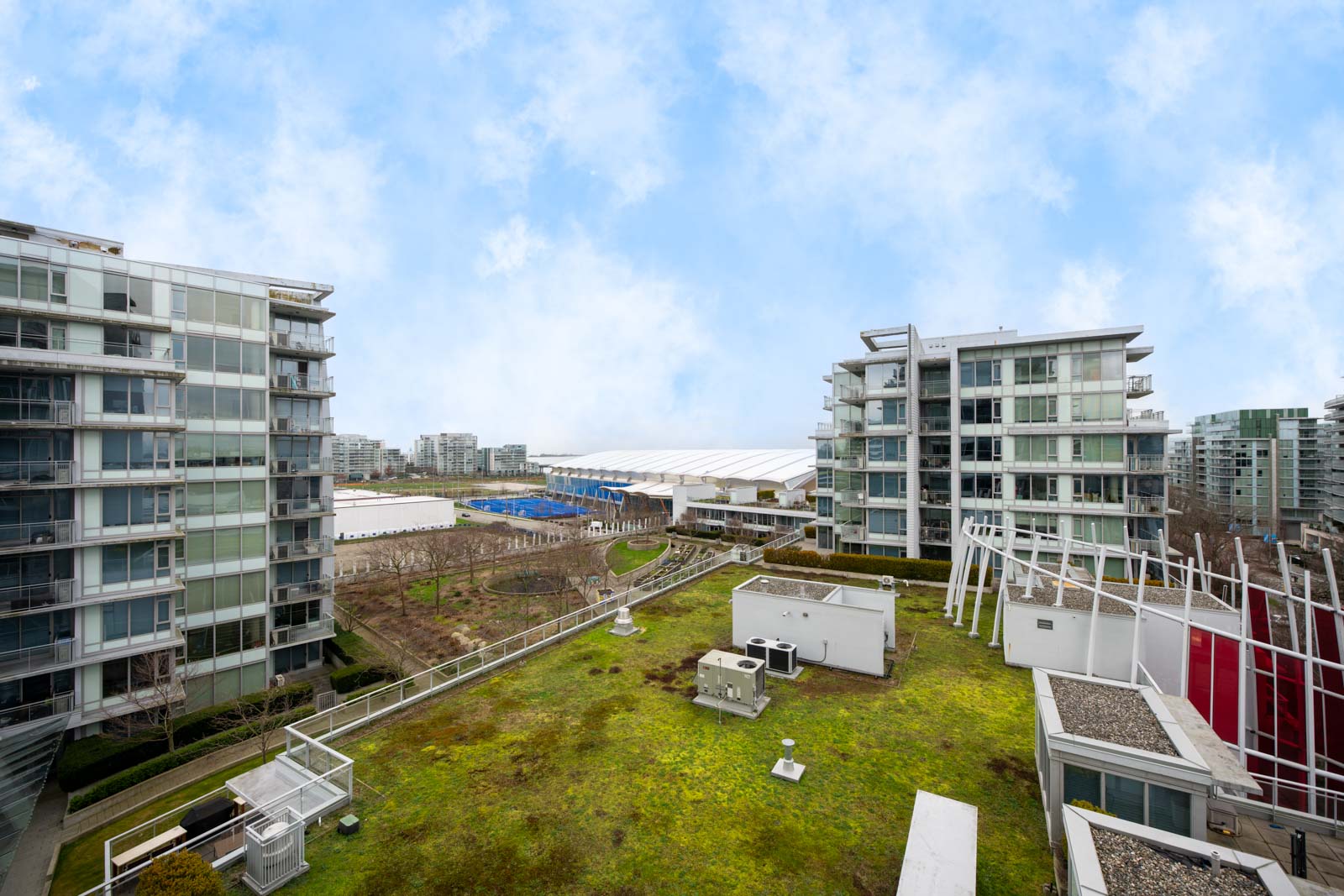 View of modern apartment buildings with glass balconies, a green rooftop with HVAC units, and a partly cloudy sky in the background.