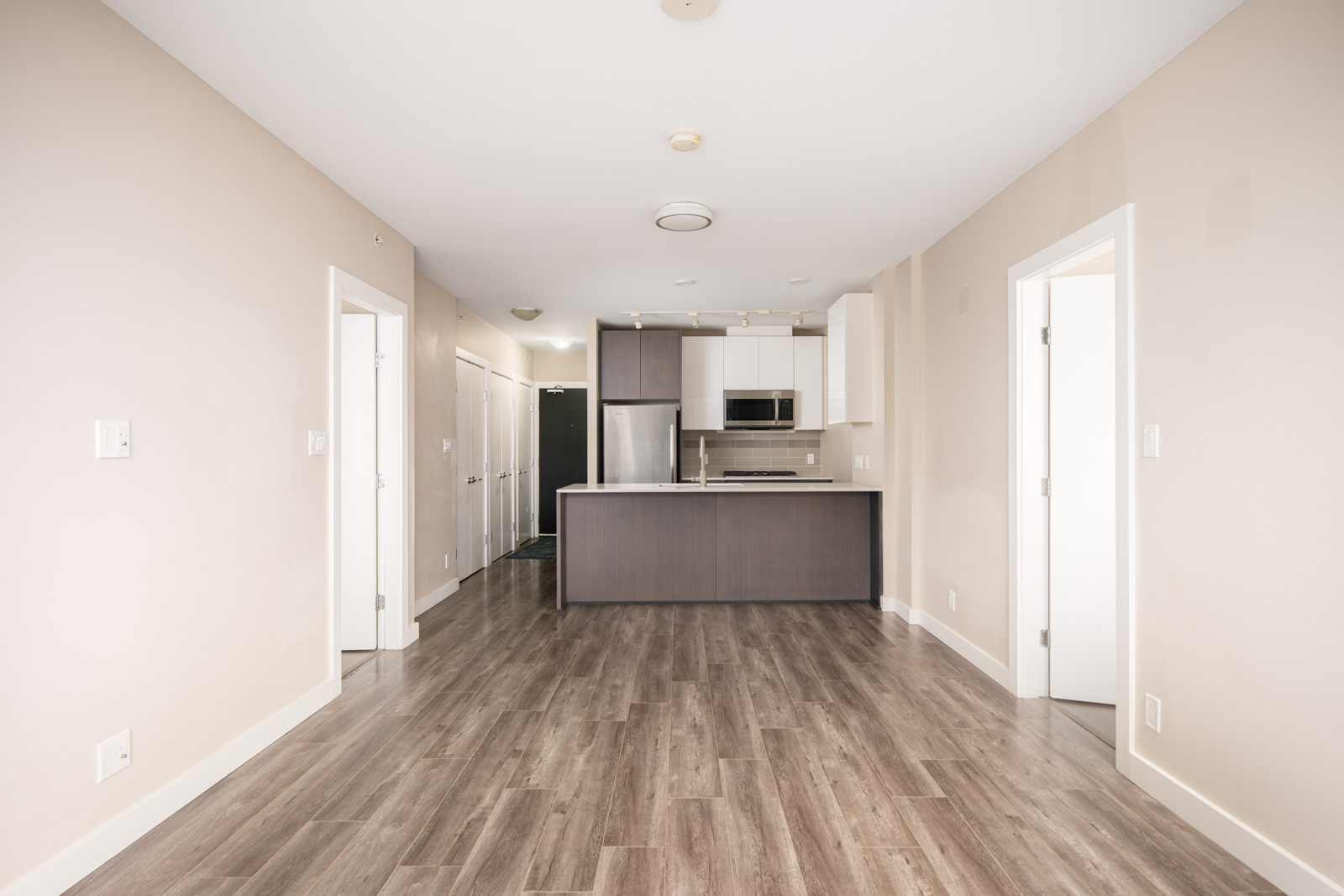 Unfurnished modern apartment with wood flooring, beige walls, an open kitchen with dark cabinets, and two doorways leading to separate rooms.
