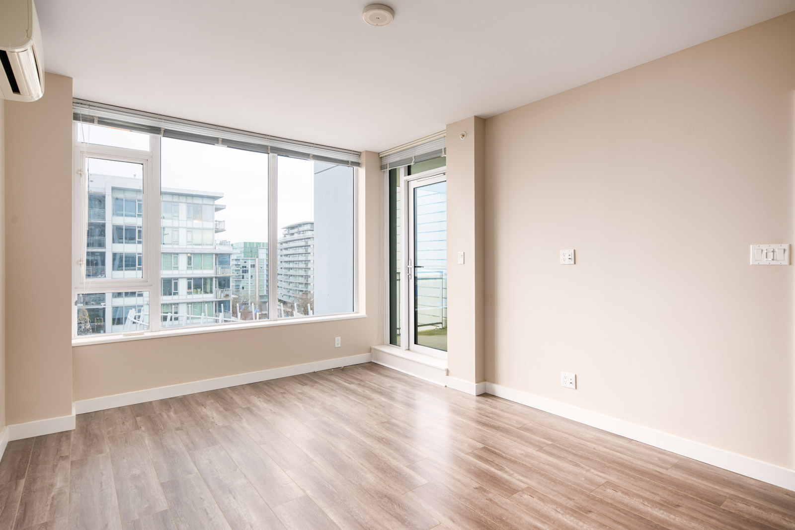 Empty modern apartment room with large windows, light-colored walls, and wood flooring overlooking nearby buildings.