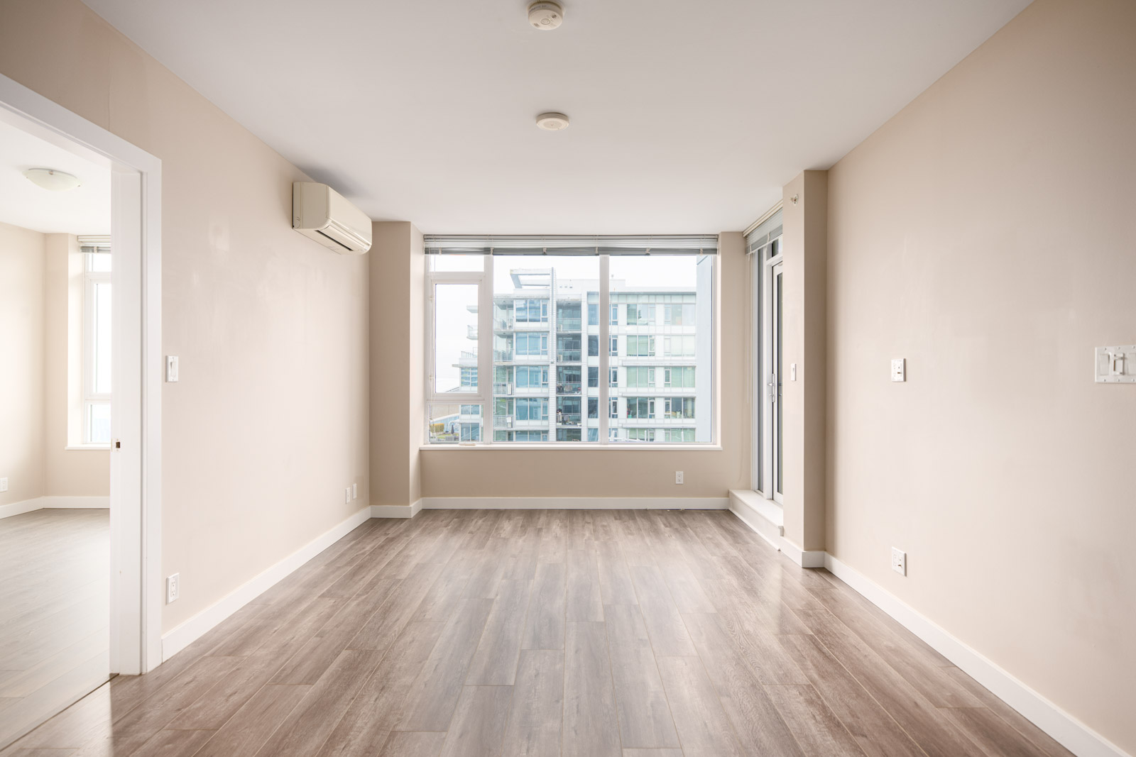 Empty apartment room with light wood flooring, beige walls, large windows showing a modern building outside, and a wall-mounted air conditioning unit.