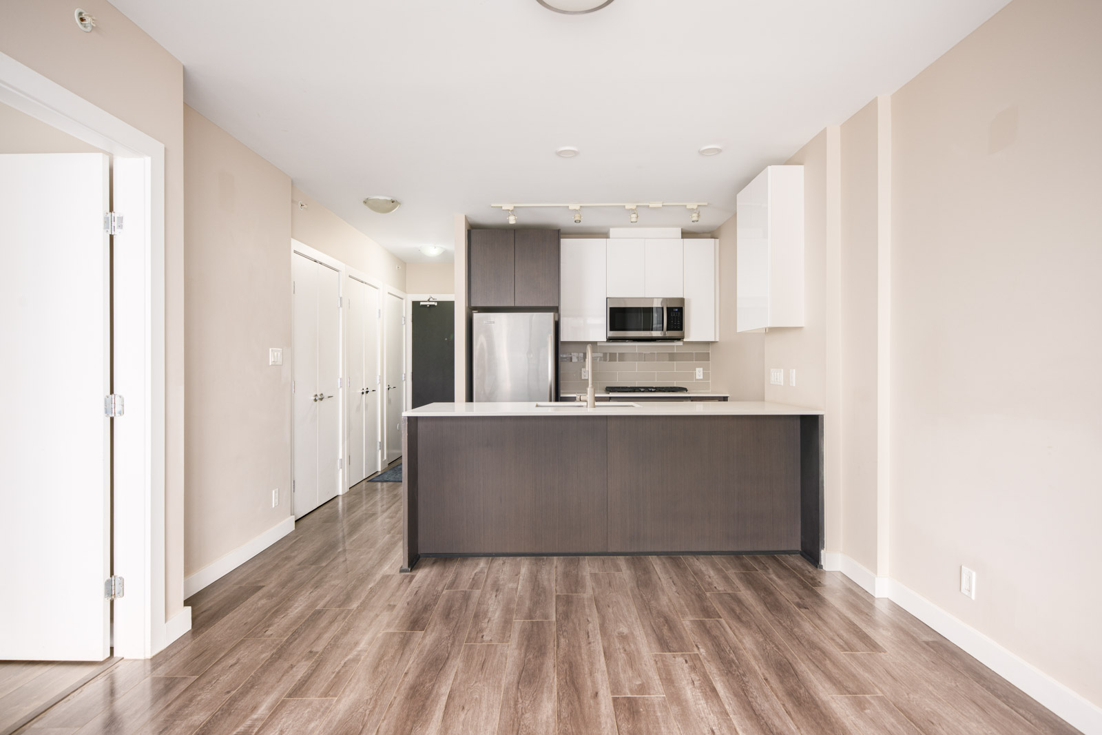 Modern, unfurnished kitchen with stainless steel appliances, white and dark cabinets, wood-look flooring, and an island counter in an open-plan apartment.