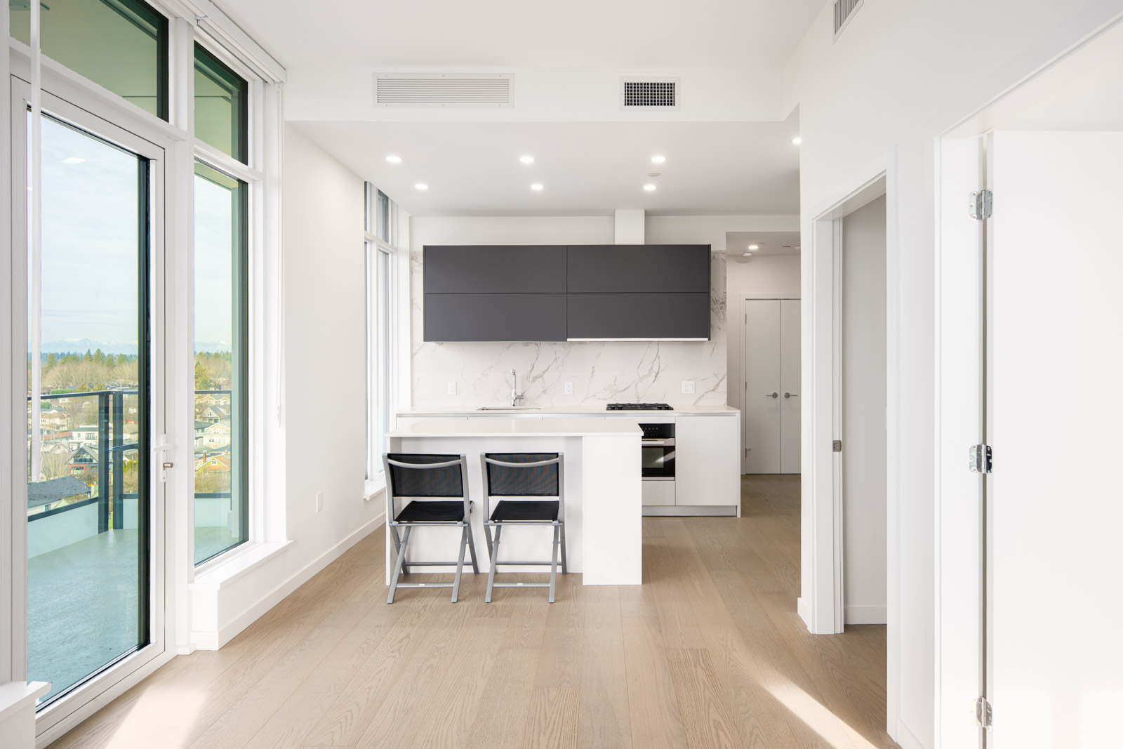 Modern apartment kitchen with white cabinets, black upper cabinets, two chairs at a breakfast bar, wood flooring, large windows, and a balcony with a city view.