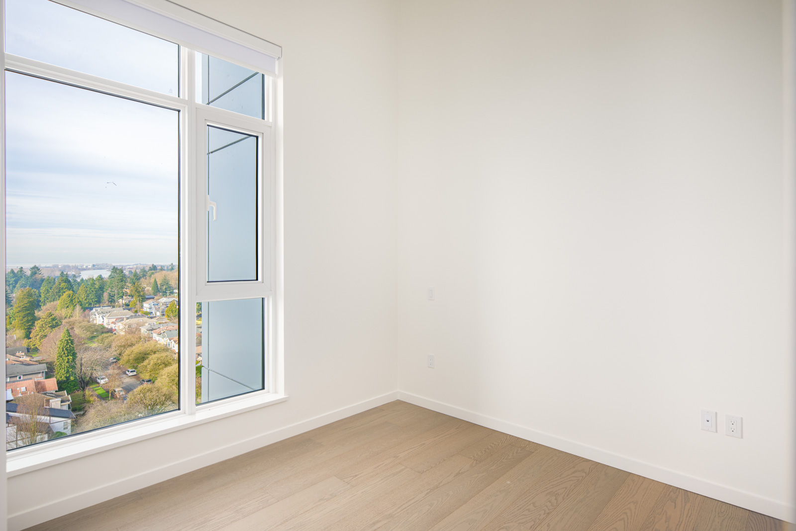 Empty room with light-colored walls and wood flooring, featuring a large window overlooking a suburban neighborhood and greenery.