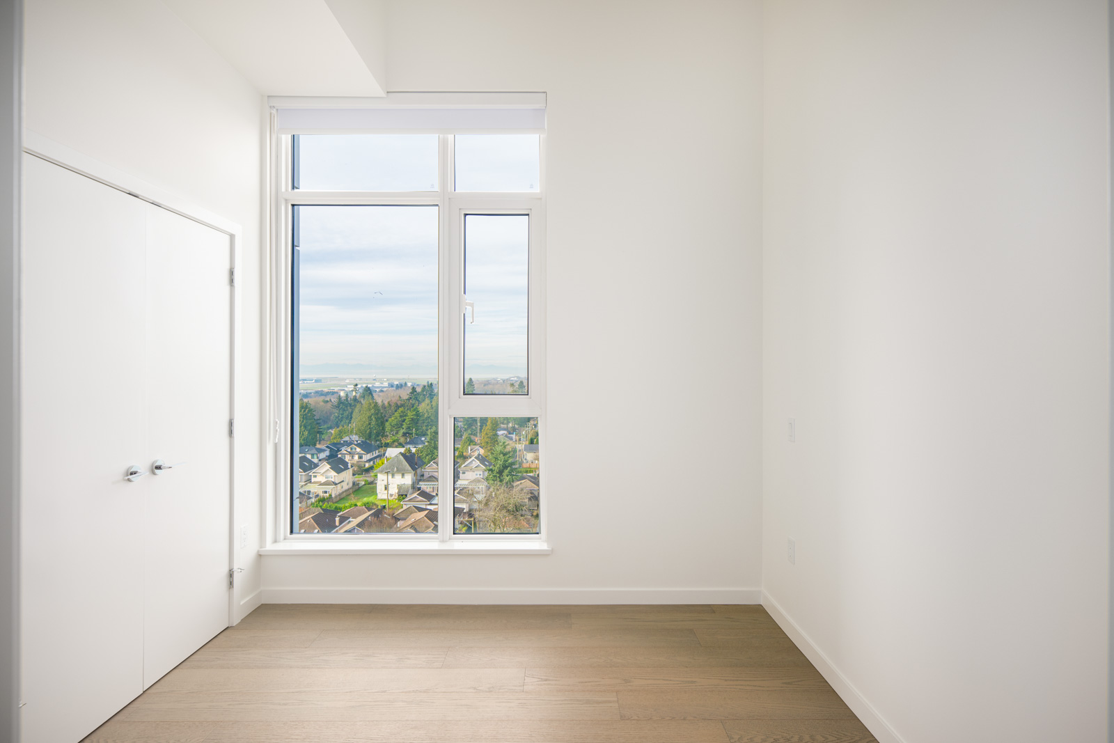 Empty room with white walls, wooden floor, double-door closet, and large window overlooking a suburban area and distant water views.
