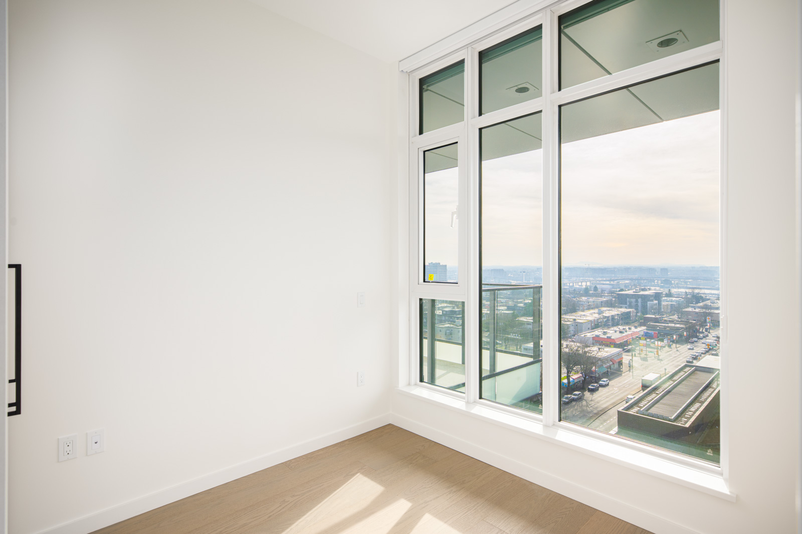 Empty corner of a room with light-colored walls, large windows, and a view overlooking a cityscape under a partly cloudy sky.