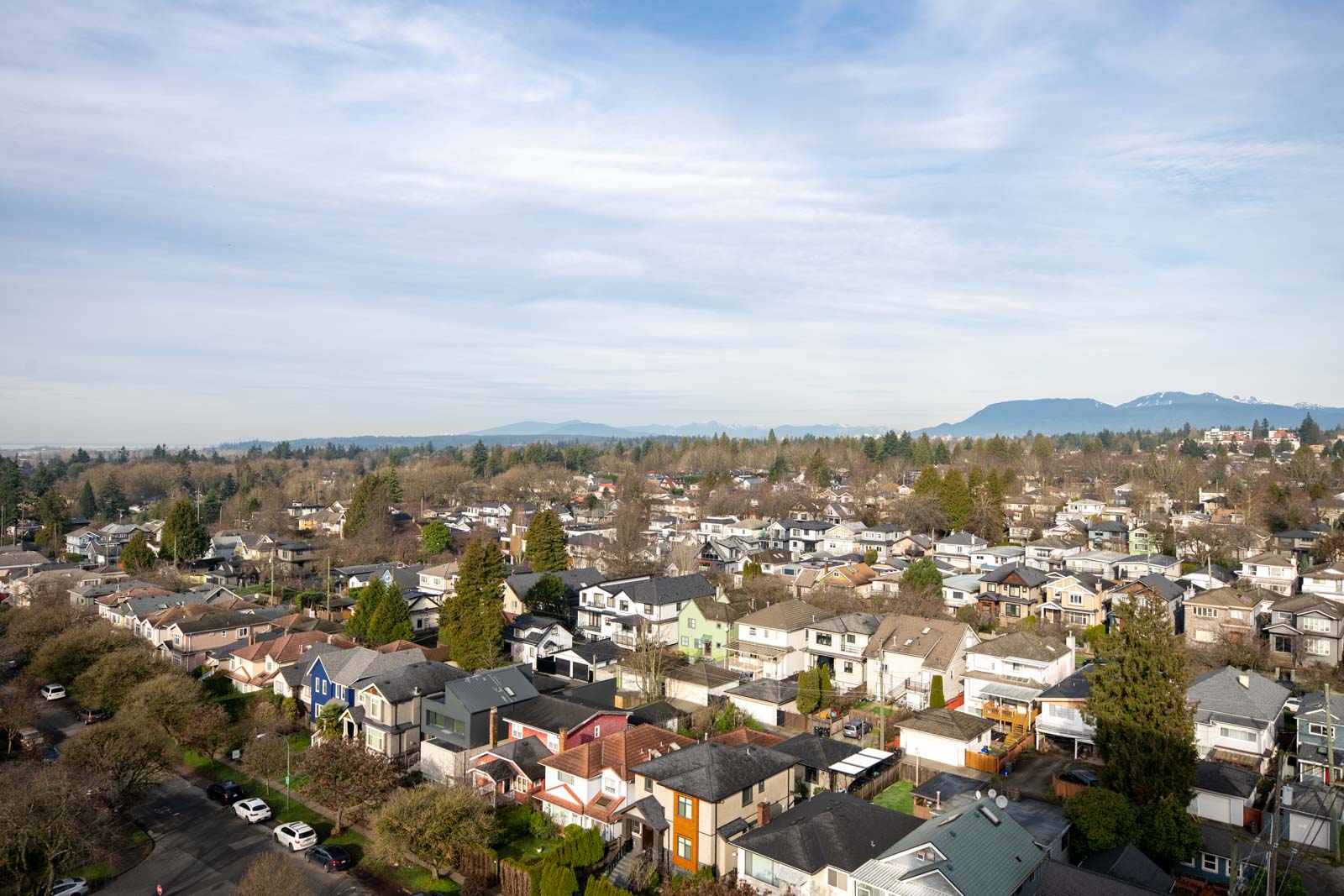 Aerial view of a suburban neighborhood with rows of houses, trees, and mountains visible in the background under a partly cloudy sky.