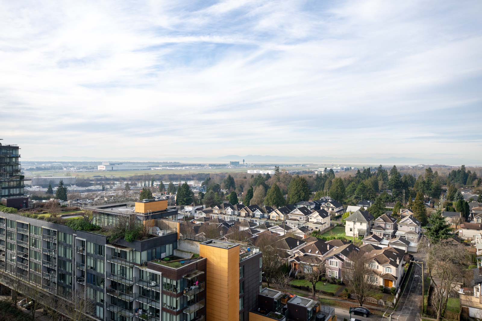 Aerial view of a residential neighborhood with houses, mid-rise buildings, trees, and a distant landscape under a partly cloudy sky.