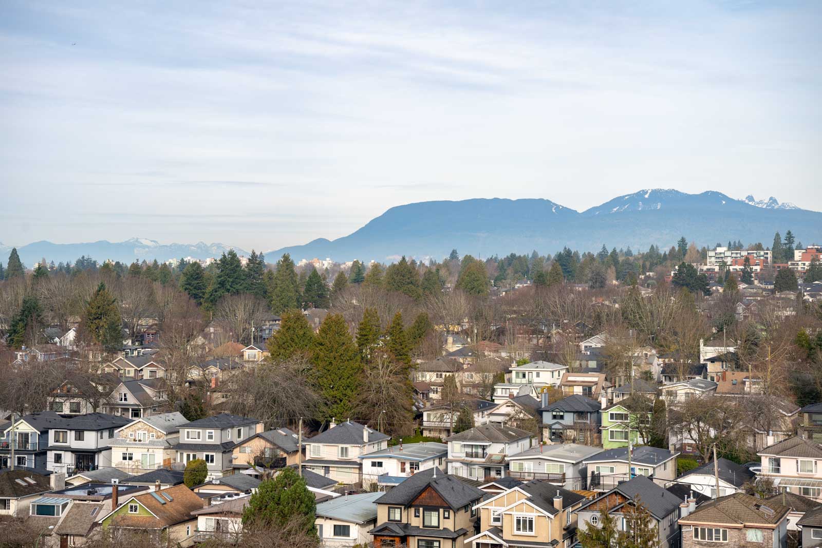 A suburban neighborhood with rows of houses, trees, and a mountain range in the background under a partly cloudy sky.