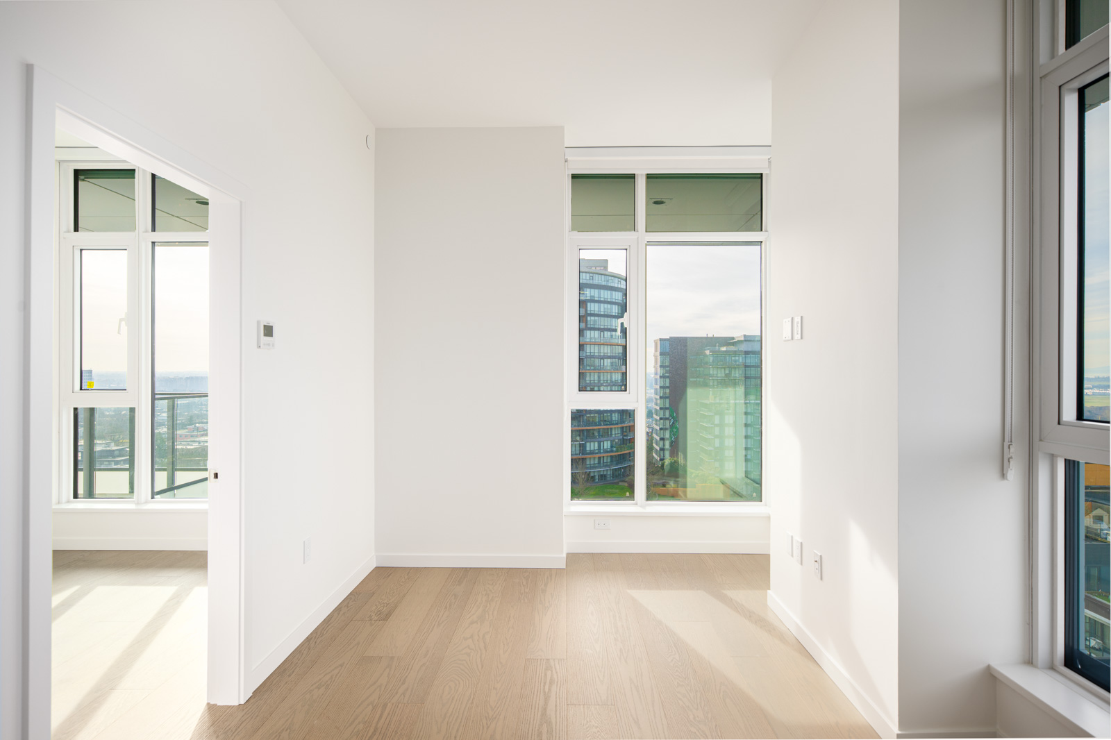 Bright, empty modern apartment room with large windows, light wood floors, and views of city buildings and a balcony.