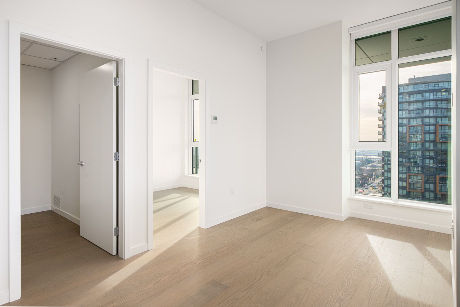 Bright, empty room with light wood flooring, two open doorways, white walls, and large windows showing a cityscape and tall building outside.