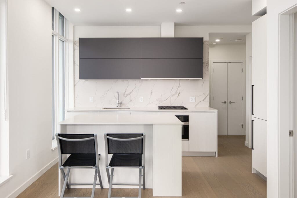 Modern kitchen with white cabinets, a marble backsplash, two black chairs at a white island, and matte grey upper cabinets. Natural light comes through tall windows on the left.