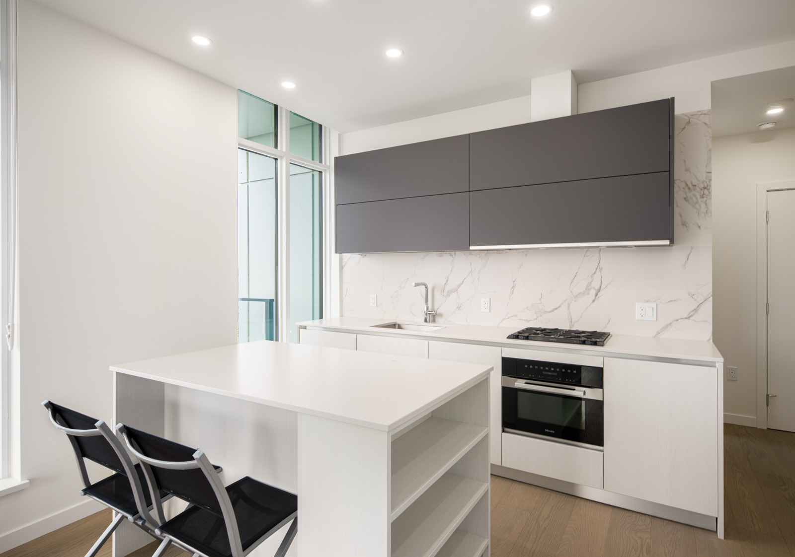 Modern kitchen with white cabinets, marble backsplash, gray upper cabinets, built-in oven, gas stove, and a white island with two black chairs.
