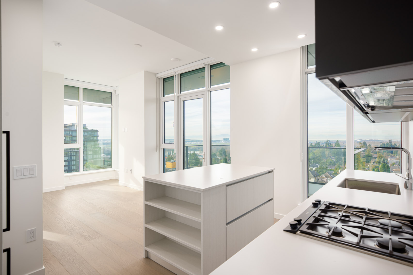 Modern, unfurnished apartment kitchen with white cabinets, gas stove, island, large windows, and a balcony offering city and distant water views in bright natural light.
